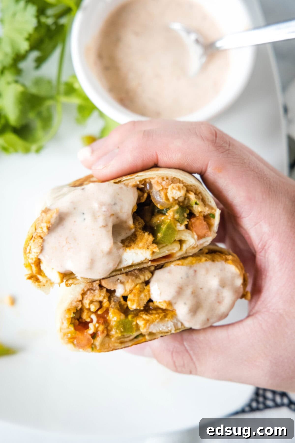 Easy chicken fajita wraps have juicy meat, tender veggies, spicy and smoky seasonings, rich beans, shredded cheese, and fresh salsa. A hand holding two halves of a fajita wrap covered in a Mexican cream, with a bowl of the sauce in the background.