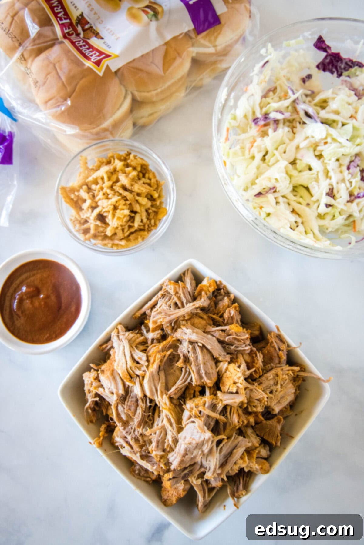 These easy pulled pork sliders combine tender and juicy meat, smoky BBQ sauce, and fresh, crunchy, tangy coleslaw. An overhead shot showcasing all the fresh ingredients needed to prepare the mouth-watering pulled pork sliders: a bowl of perfectly shredded pulled pork, a bowl of creamy coleslaw, a generous serving of barbecue sauce, crispy fried onions, and a bag of soft slider buns.