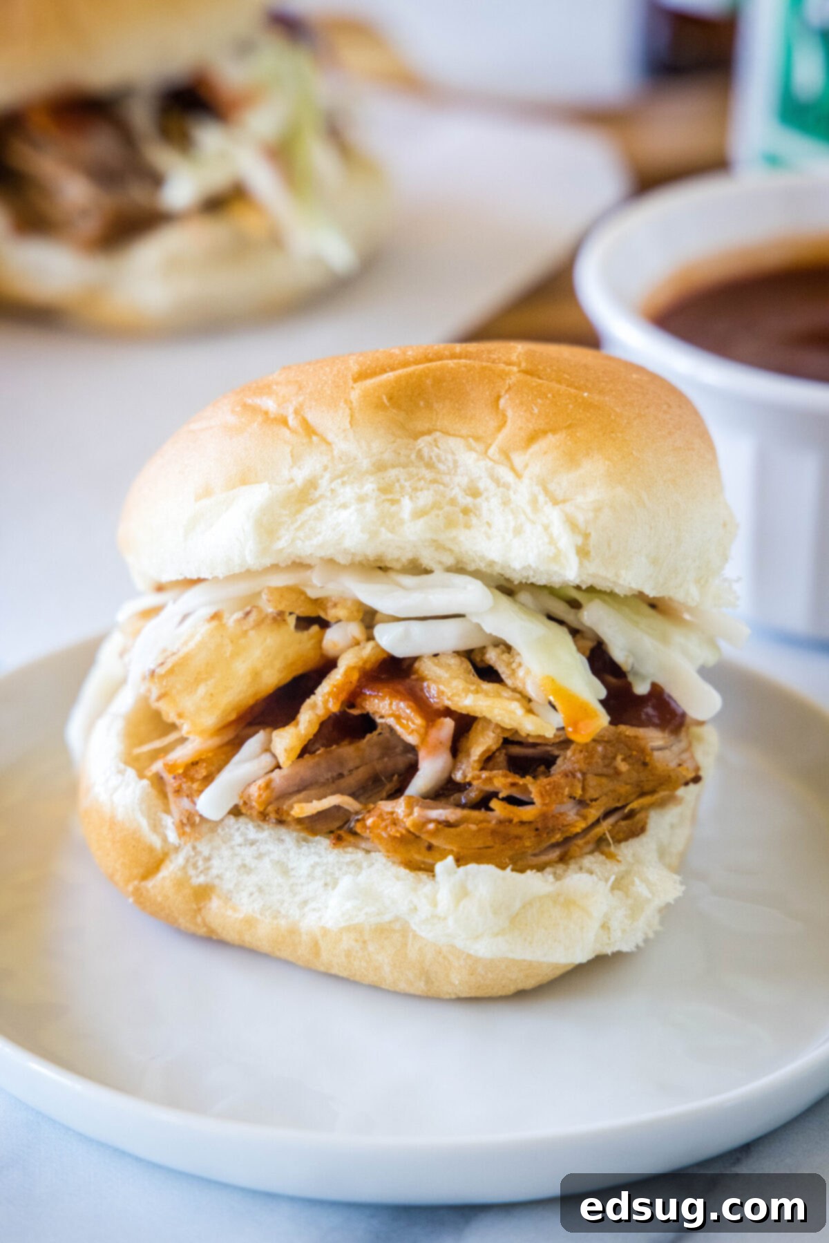 These easy pulled pork sliders deliver tender and juicy meat, topped with smoky BBQ sauce, and fresh, crunchy, tangy coleslaw. A perfectly assembled pulled pork slider with visible coleslaw nestled on a white plate, with another slider enticingly blurred in the background, highlighting its deliciousness.