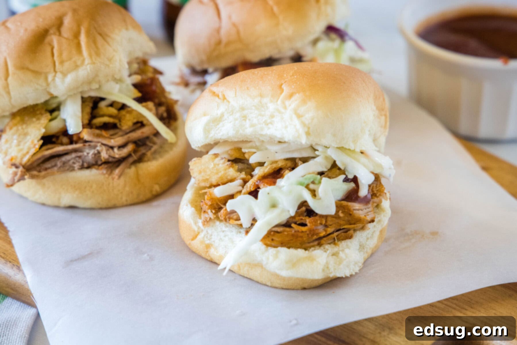 A trio of easy pulled pork sliders with tender and juicy meat, topped with smoky BBQ sauce, and fresh, crunchy, tangy coleslaw. Three perfectly plated pulled pork sliders sitting next to a small bowl of rich barbecue sauce, ready for serving.