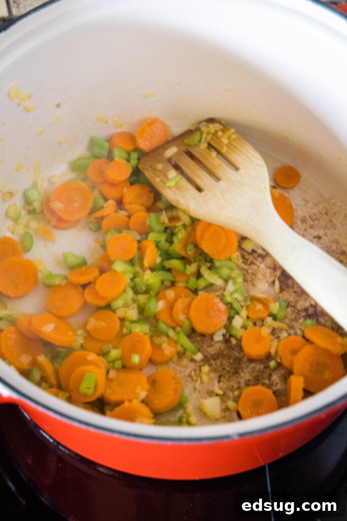 Sautéing Mirepoix for Flavorful Chicken Rice Soup. Diced carrots, onion, and celery, the aromatic base, sautéing in a large pot with cooking oil, stirred by a wooden spatula.