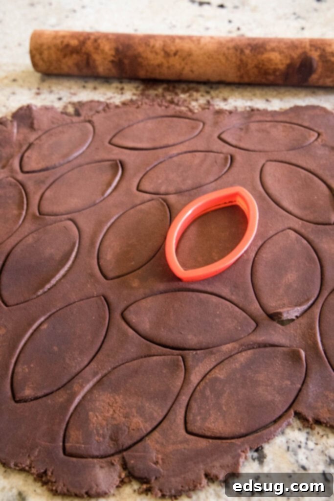 A red cookie cutter is used to cut chocolate sugar cookie dough into football shapes on a cocoa-dusted surface.