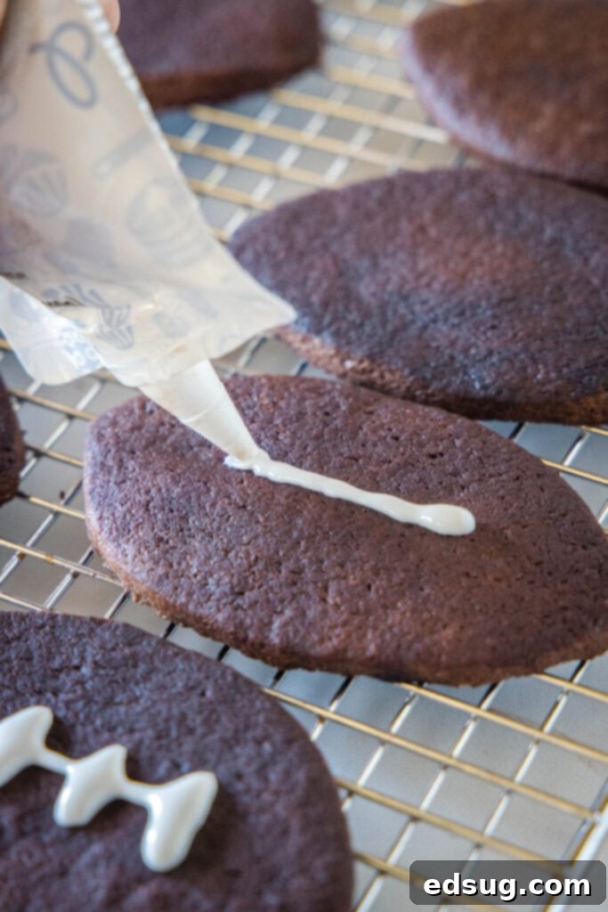 A piping tip applies white lines of frosting over football-shaped chocolate sugar cookies, creating laces on a wire rack.