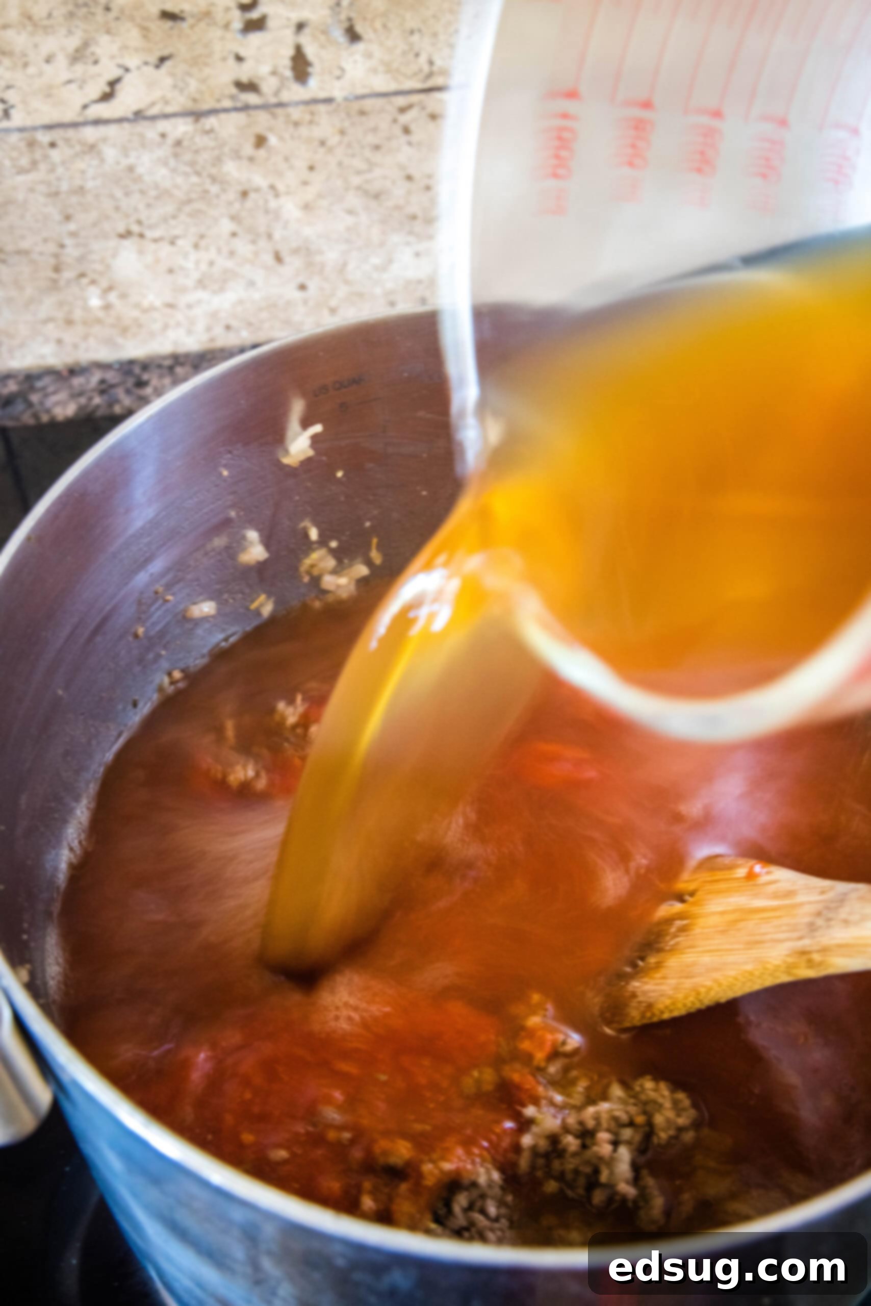 A large pot on the stovetop filled with beef macaroni soup ingredients, with beef broth being poured in, illustrating the liquid base being added.