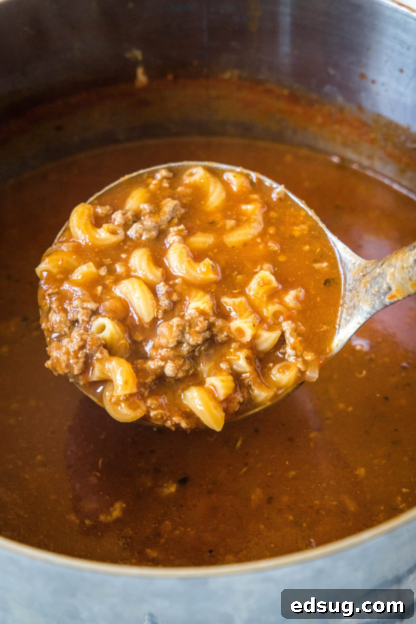 A ladle scooping a generous portion of steaming beef macaroni soup from a large pot, showcasing the rich broth, ground beef, and macaroni.