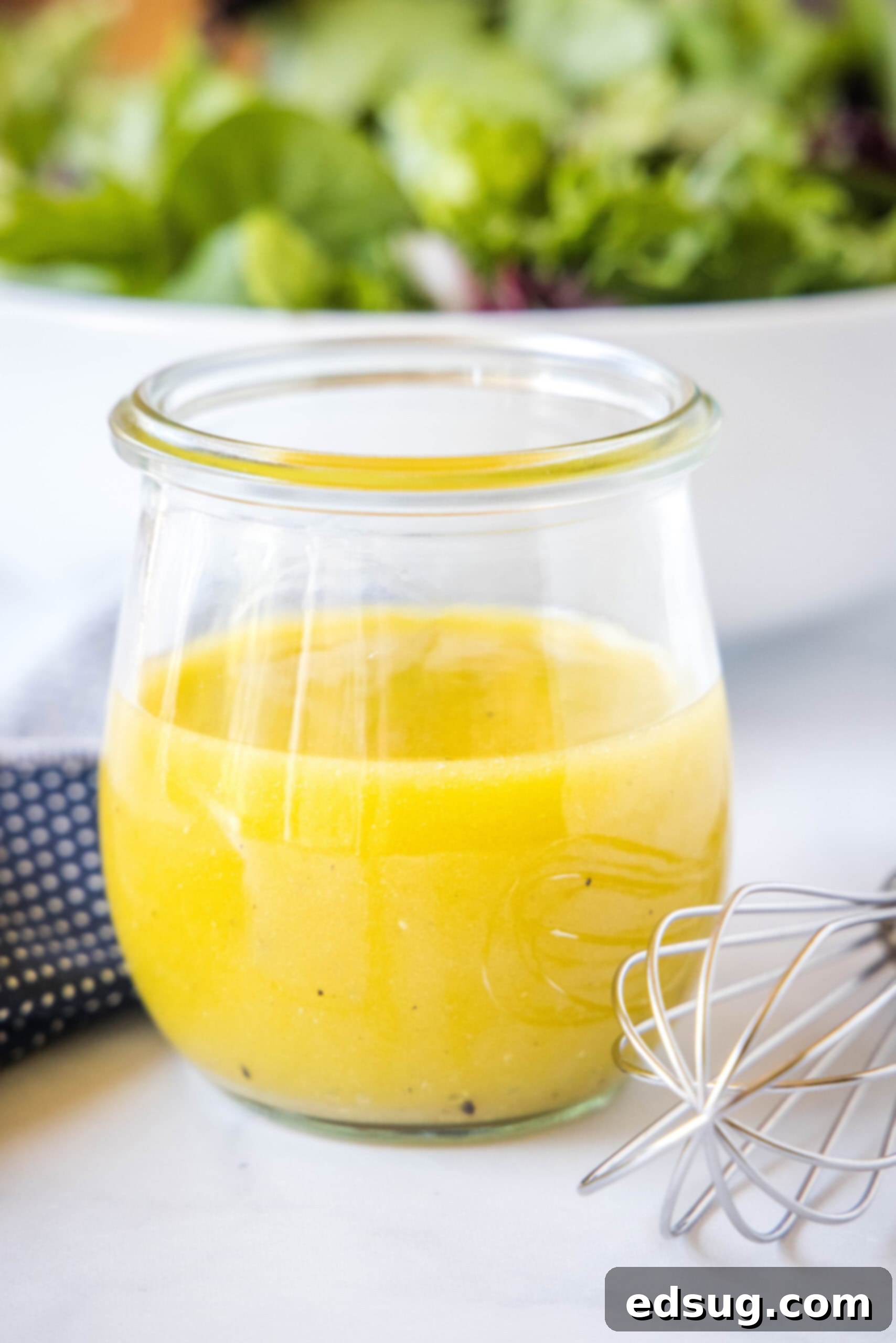 A jar of homemade apple cider vinaigrette on a countertop next to a whisk, with a bowl of fresh fall salad in the background.