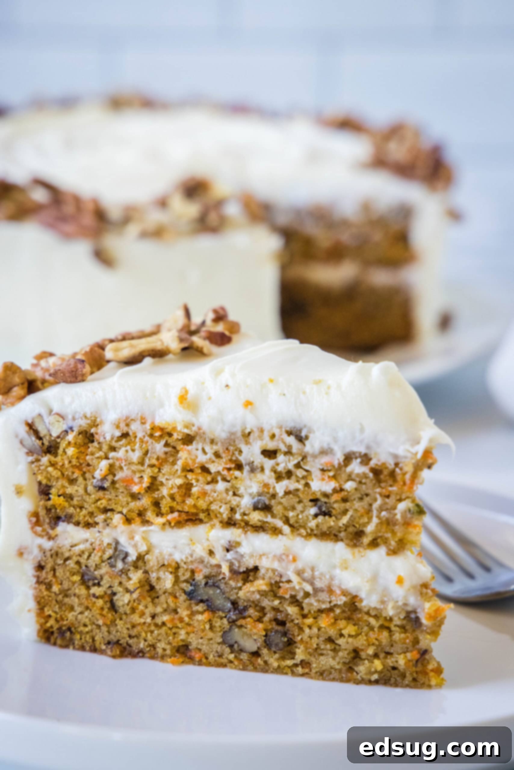 The best homemade carrot cake! Made from moist cake layers loaded with crunchy pecans, filled and frosted with sweet cream cheese frosting. A slice of frosted carrot cake on a white plate next to a fork, with the rest of the cake in the background.