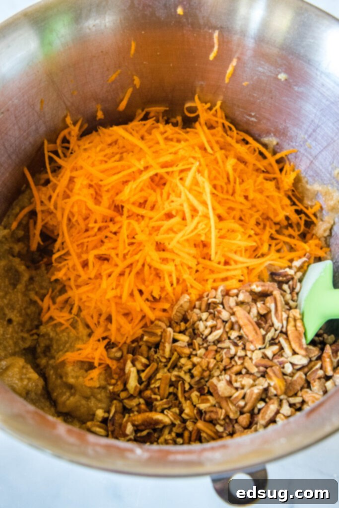 The best homemade carrot cake! Made from moist cake layers loaded with crunchy pecans, filled and frosted with sweet cream cheese frosting. Shredded carrots and chopped pecans added to carrot cake batter in a metal mixing bowl.