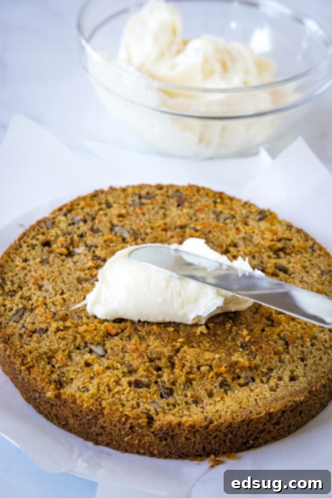 The best homemade carrot cake! Made from moist cake layers loaded with crunchy pecans, filled and frosted with sweet cream cheese frosting. An offset spatula spreading cream cheese frosting over a carrot cake layer, with a glass bowl of frosting in the background.