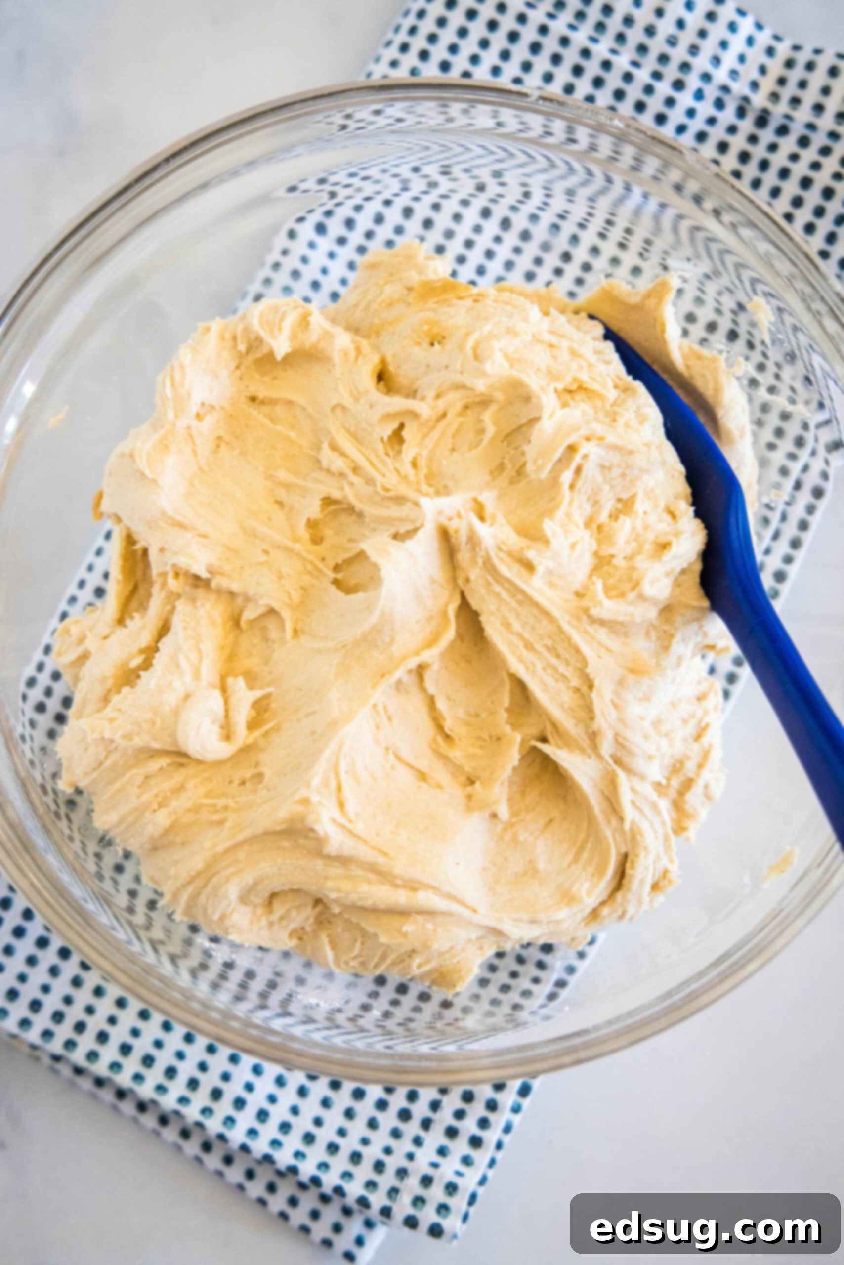 Homemade peanut butter frosting is smooth, creamy, and perfect for decorating cakes and desserts. Ready in minutes with just 5 ingredients! Overhead view of peanut butter frosting in a glass bowl with a blue stirring spoon.