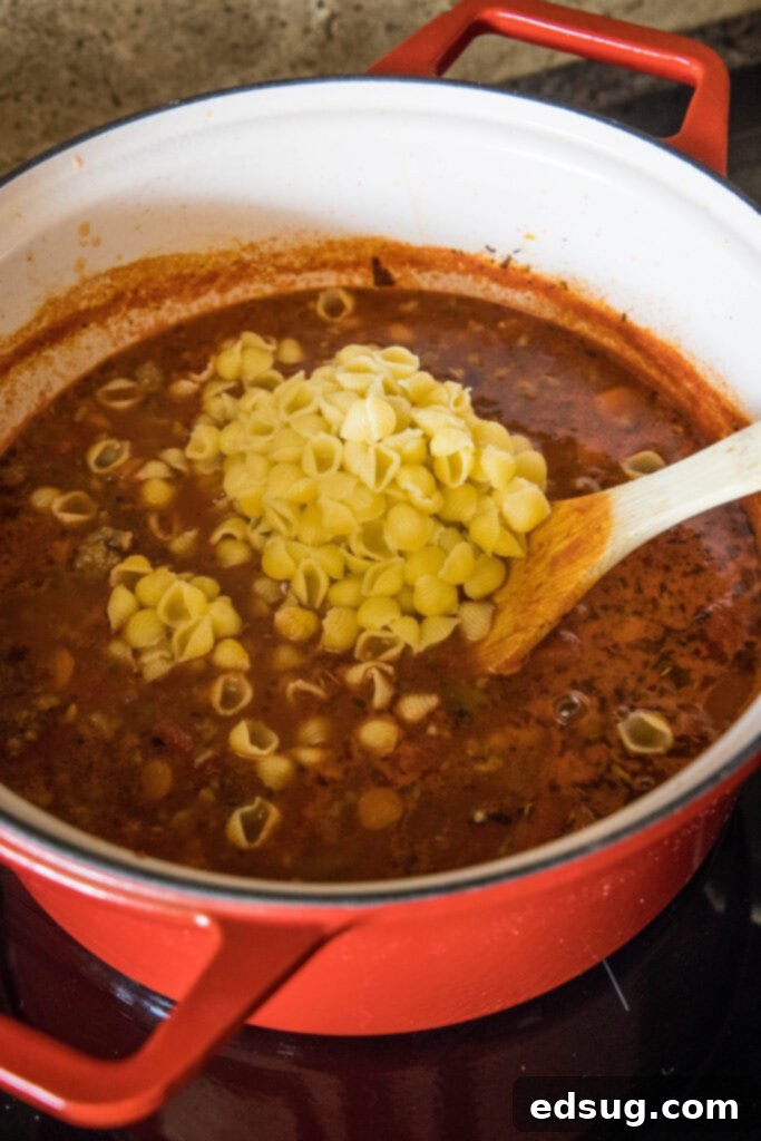 Uncooked small pasta shells being added to a pot of simmering Italian sausage soup.