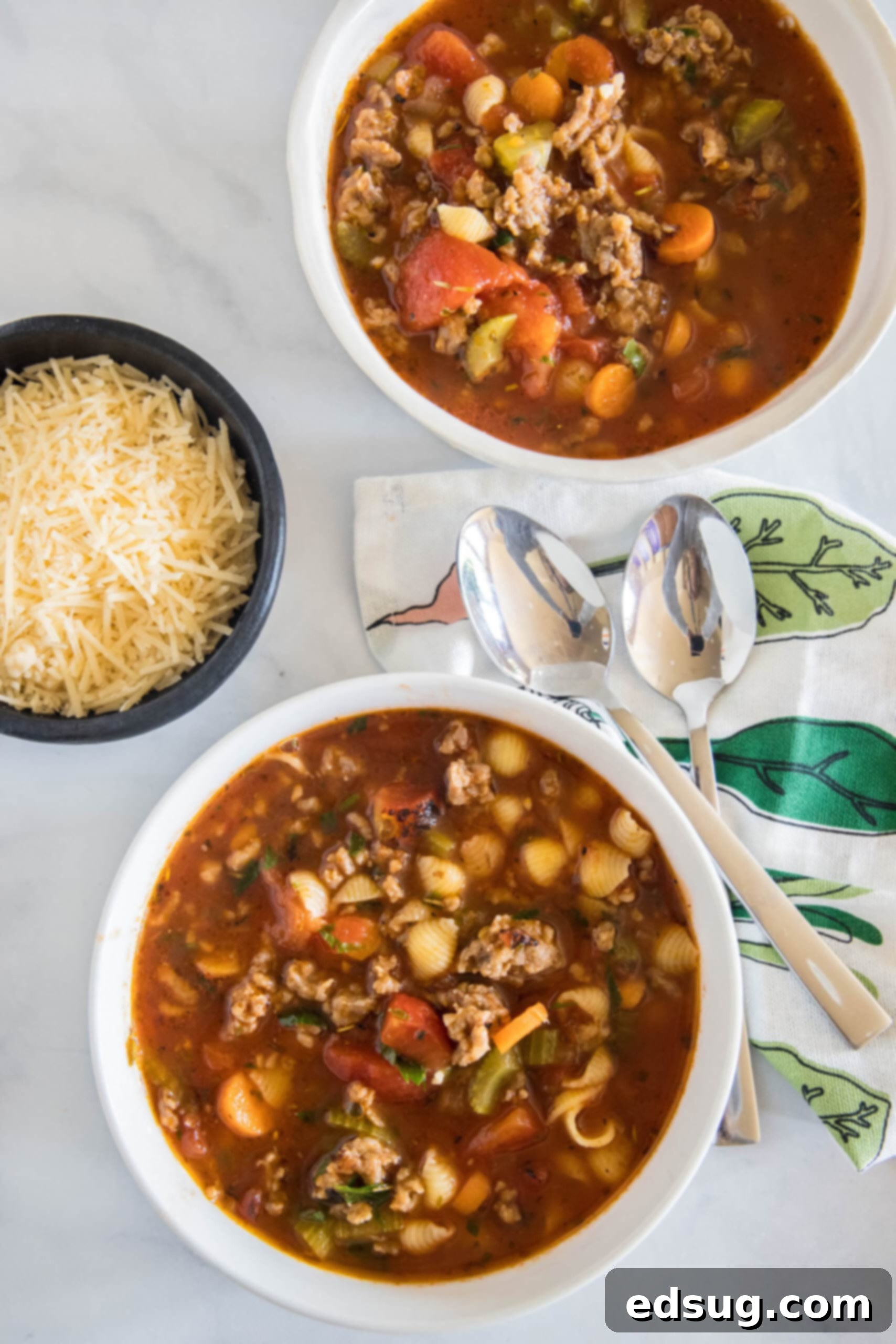 Two bowls of Italian sausage soup, generously topped with shredded cheese, on a kitchen counter with spoons nearby.