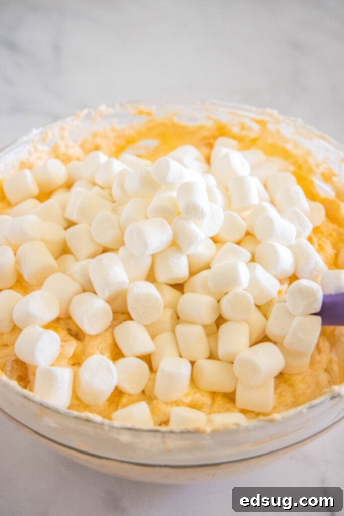 Marshmallows added to orange fluff in a glass bowl.