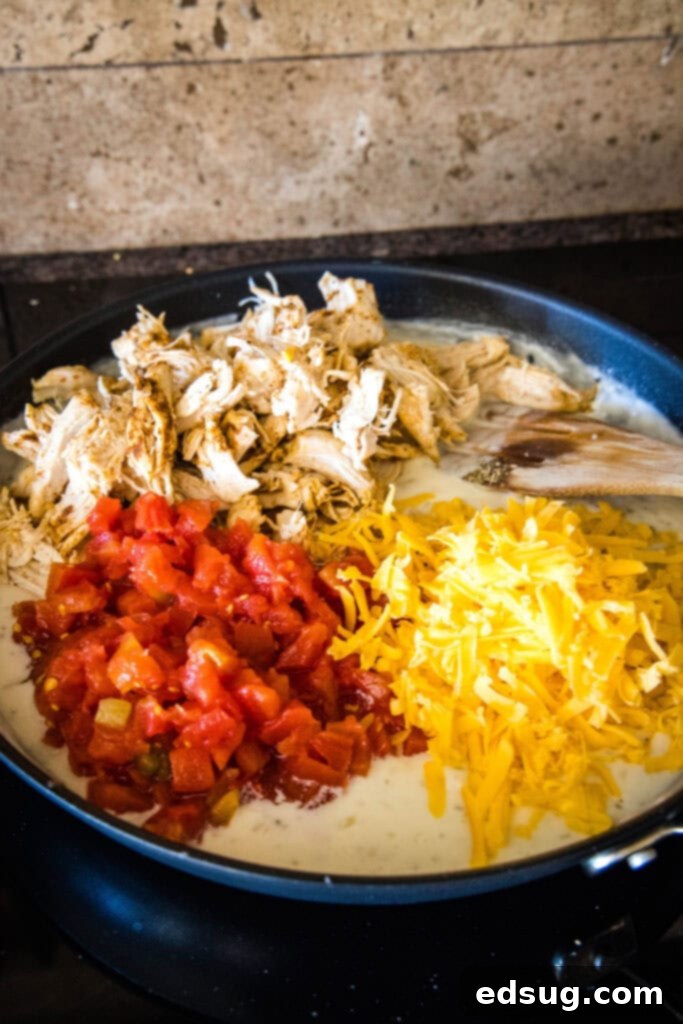 Make this casserole-style cheesy chicken spaghetti with Rotel tomatoes, juicy chicken, and tender pasta baked in a flavorful creamy sauce. Rotel tomatoes, shredded chicken, and shredded cheese added to cream sauce in a skillet, next to a wooden spoon.