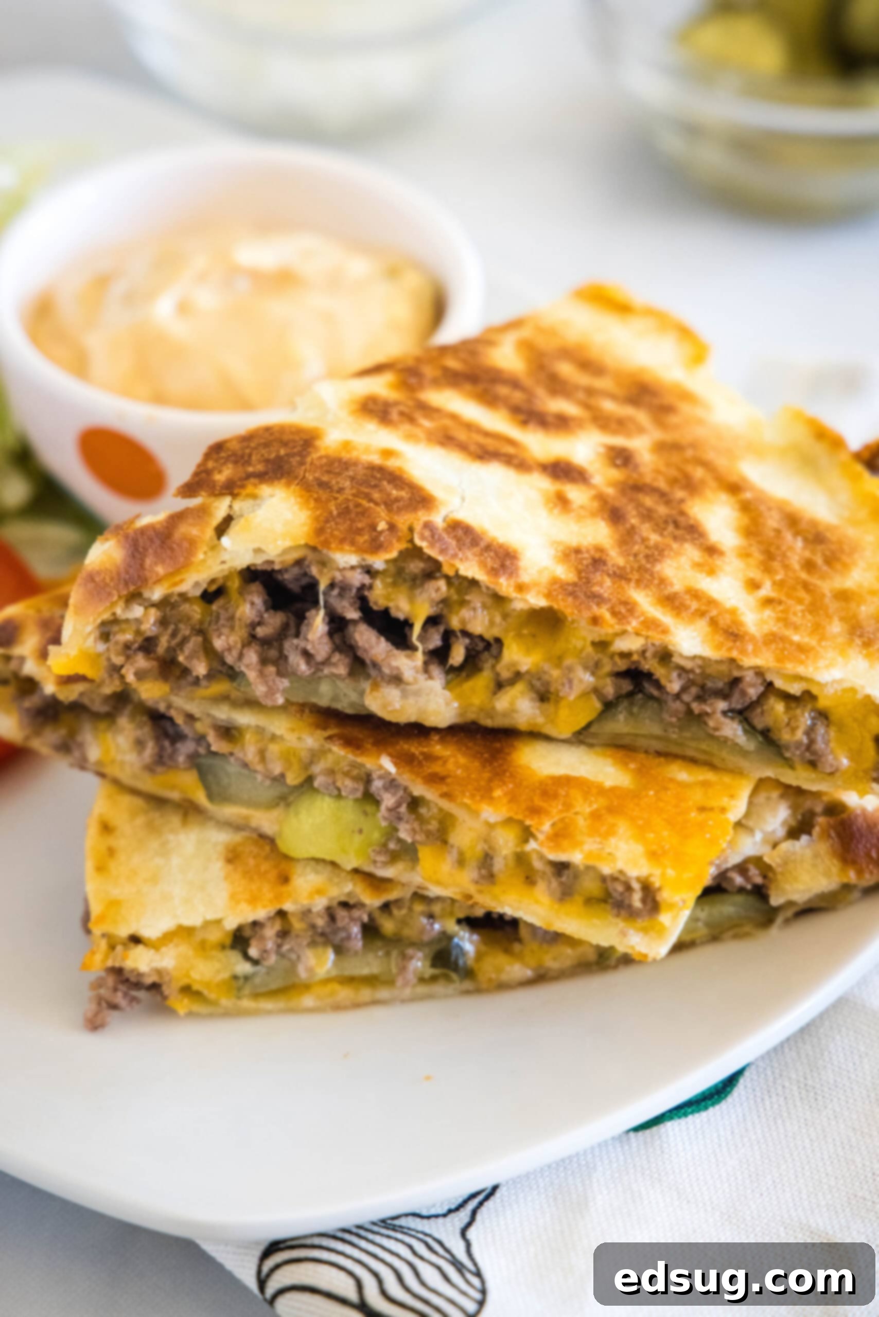 Tuck into a crispy cheeseburger quesadilla loaded with juicy burger meat, onions, pickles, and melty cheese folded inside a warm tortilla. Three slices of cheeseburger quesadilla stacked on a plate, next to a bowl of burger sauce.