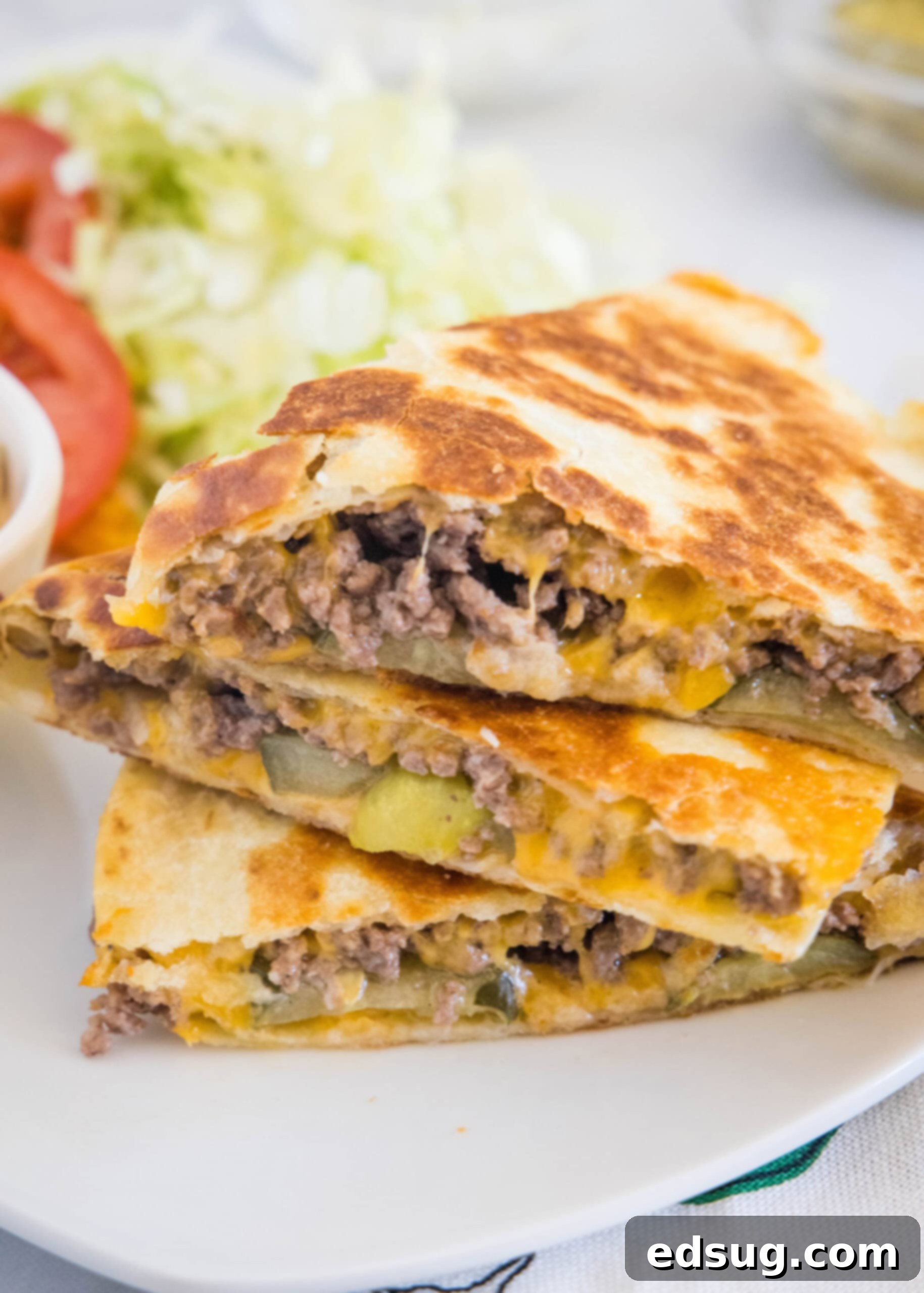 Tuck into a crispy cheeseburger quesadilla loaded with juicy burger meat, onions, pickles, and melty cheese folded inside a warm tortilla. Three slices of cheeseburger quesadilla stacked on a plate, with lettuce and sliced tomatoes in the background.