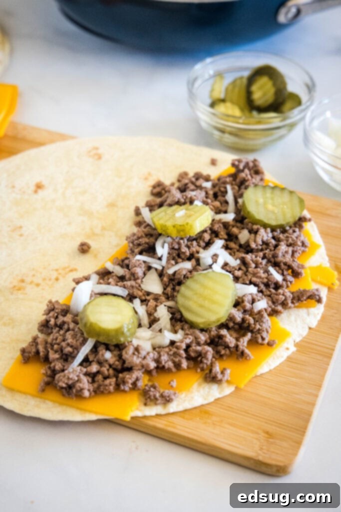 Tuck into a crispy cheeseburger quesadilla loaded with juicy burger meat, onions, pickles, and melty cheese folded inside a warm tortilla. A flour tortilla laying open on a wooden board, one half topped with cheese, burger meat, onions, and pickles.