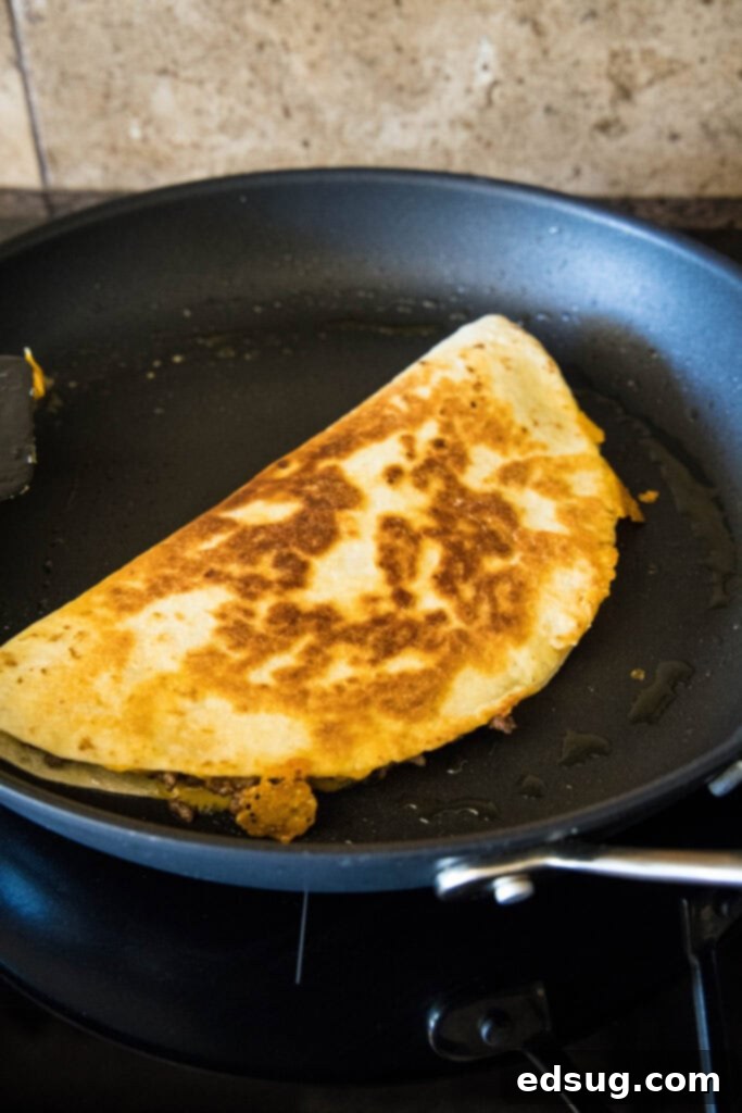 Tuck into a crispy cheeseburger quesadilla loaded with juicy burger meat, onions, pickles, and melty cheese folded inside a warm tortilla. A cheeseburger quesadilla frying in a black skillet.