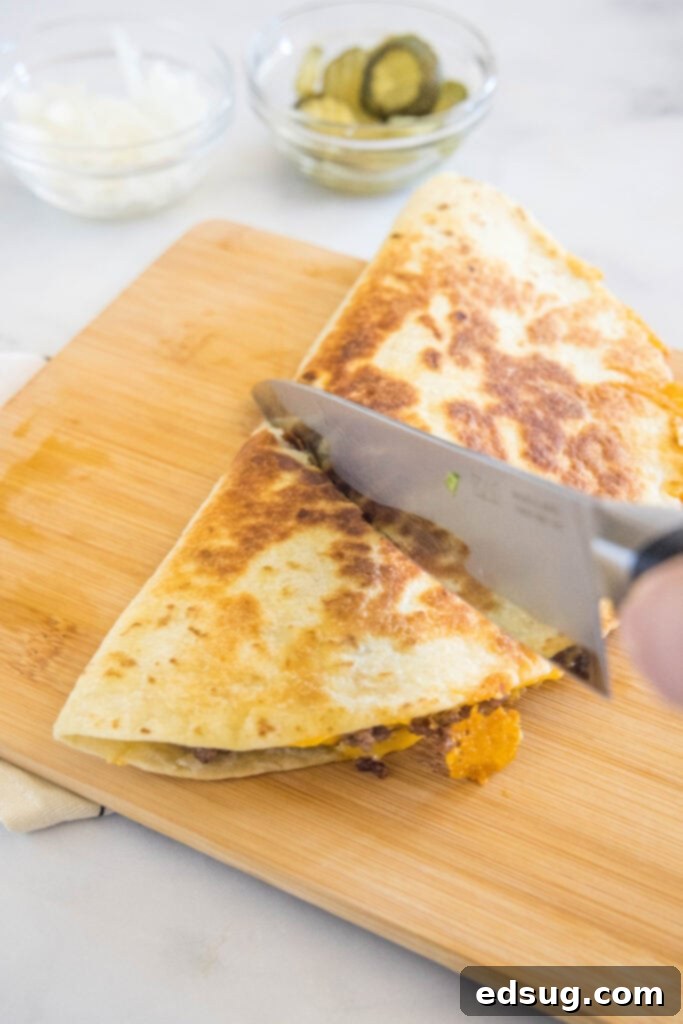 Tuck into a crispy cheeseburger quesadilla loaded with juicy burger meat, onions, pickles, and melty cheese folded inside a warm tortilla. A knife cutting a cheeseburger quesadilla into slices on a wooden cutting board.