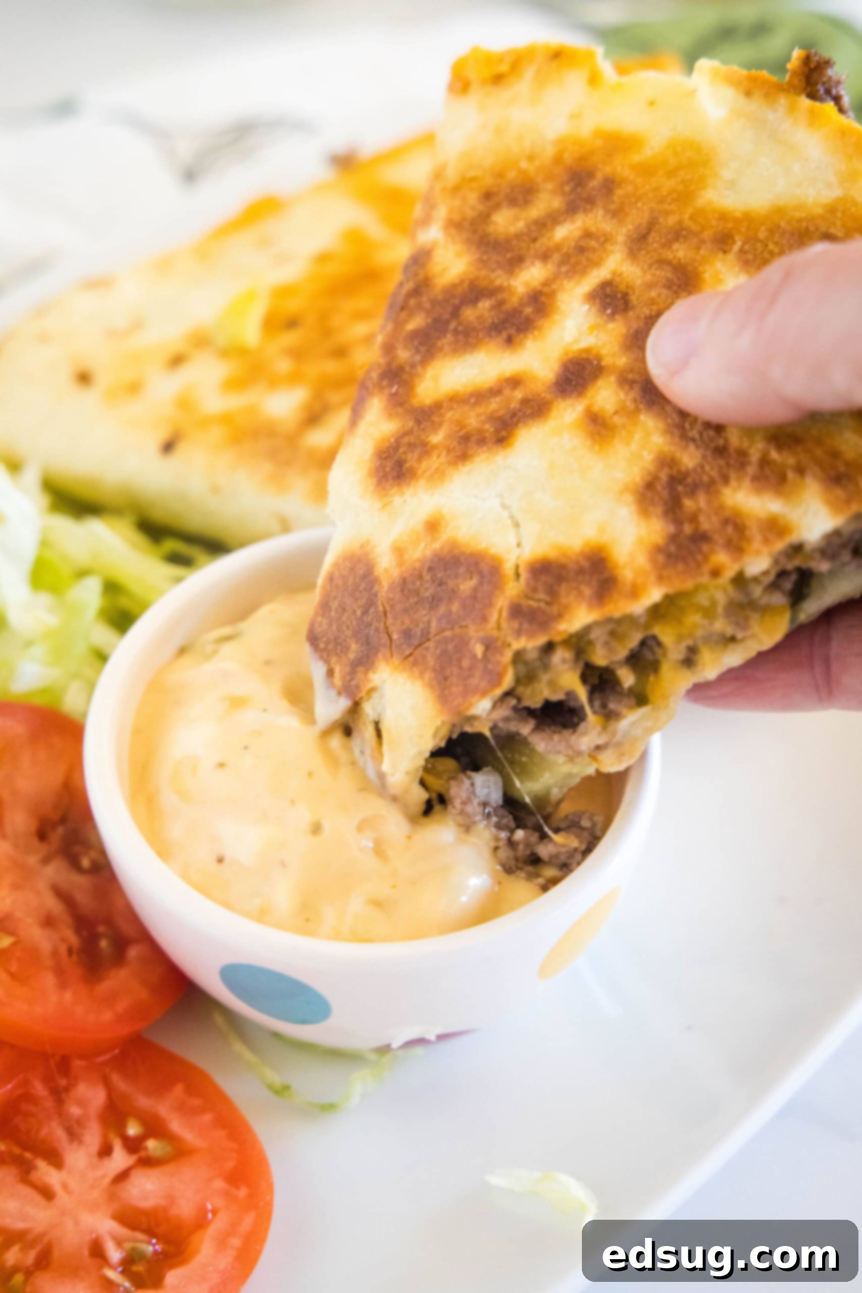 Tuck into a crispy cheeseburger quesadilla loaded with juicy burger meat, onions, pickles, and melty cheese folded inside a warm tortilla. A hand dipping a cheeseburger quesadilla into a small bowl of burger sauce, with more quesadillas in the background.