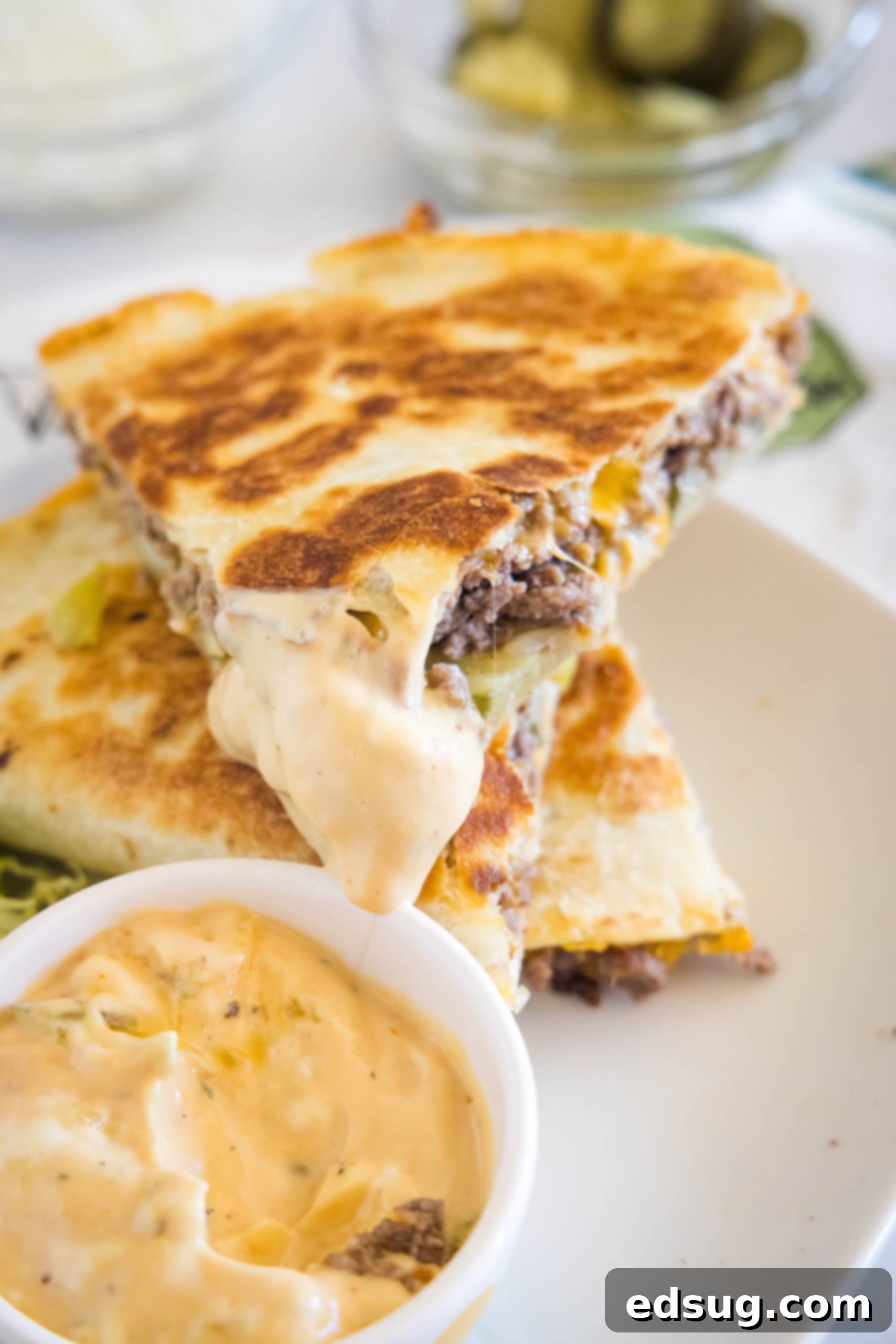 Tuck into a crispy cheeseburger quesadilla loaded with juicy burger meat, onions, pickles, and melty cheese folded inside a warm tortilla. A slice of cheeseburger quesadilla dipped into a bowl of burger sauce, stacked on top of another slice on a white plate.
