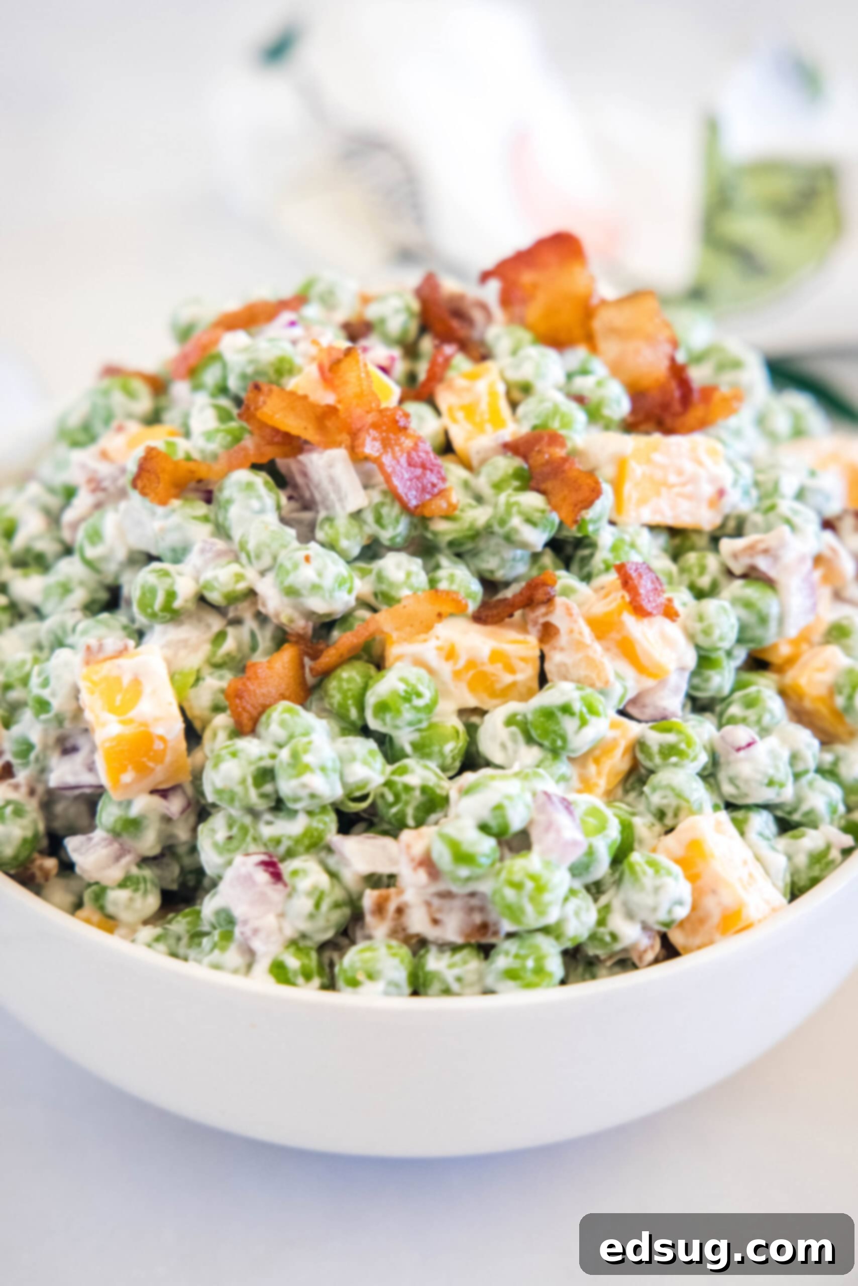 Cool and creamy pea salad is an easy side packed with crispy bacon, cheddar morsels, and red onions tossed in tangy sour cream dressing. Vibrant green pea salad in a pristine white serving bowl, garnished with fresh herbs and a sprinkle of bacon.