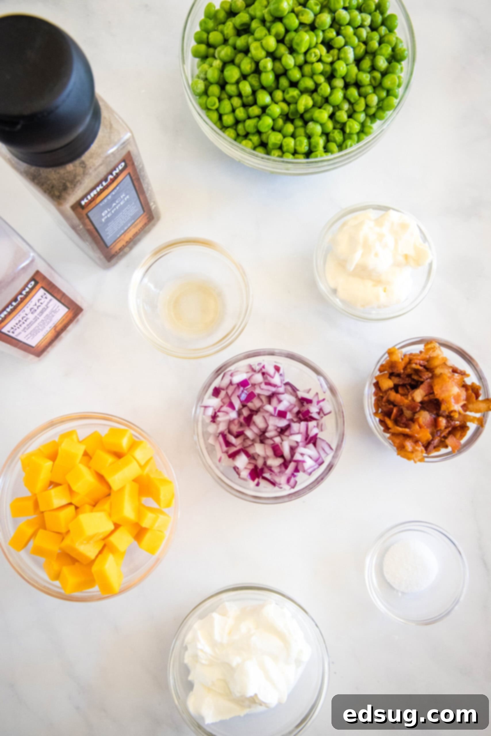 Cool and creamy pea salad is an easy side packed with crispy bacon, cheddar morsels, and red onions tossed in tangy sour cream dressing. Various fresh ingredients for pea salad laid out on a white surface, including peas, bacon, cheddar, and onion.