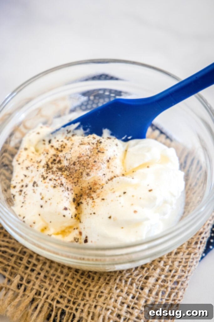 Cool and creamy pea salad is an easy side packed with crispy bacon, cheddar morsels, and red onions tossed in tangy sour cream dressing. Ingredients for the creamy dressing being whisked together in a glass bowl with a blue rubber spatula.