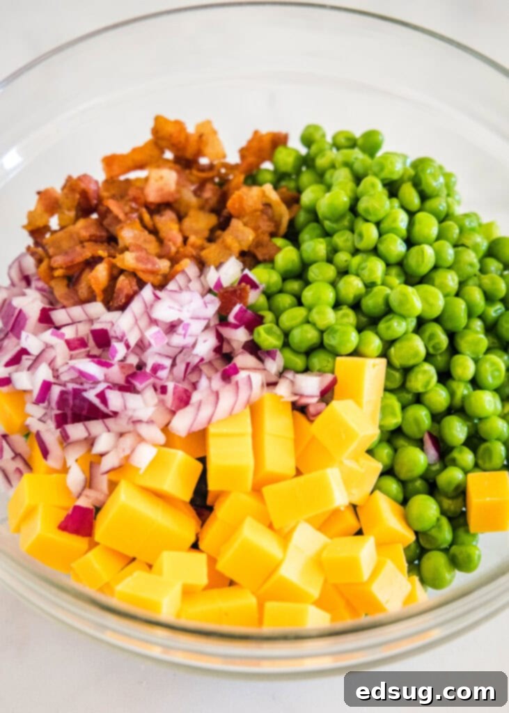 Cool and creamy pea salad is an easy side packed with crispy bacon, cheddar morsels, and red onions tossed in tangy sour cream dressing. All pea salad ingredients, including peas, bacon, cheddar, and onion, combined in a large glass mixing bowl before dressing is added.