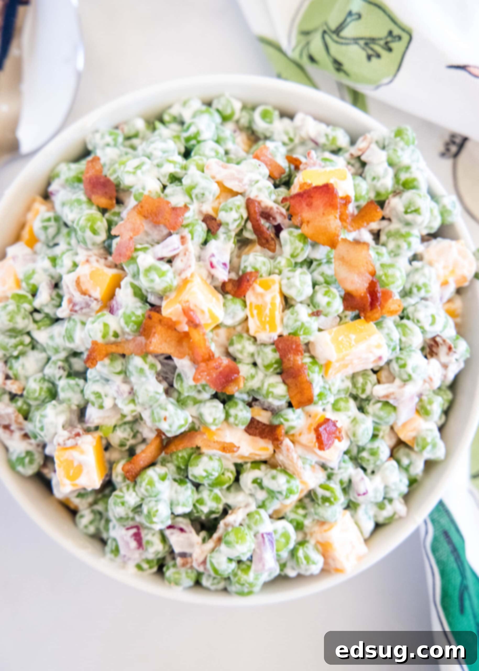 Cool and creamy pea salad is an easy side packed with crispy bacon, cheddar morsels, and red onions tossed in tangy sour cream dressing. Overhead close-up view of creamy pea salad in a white bowl, showing crispy bacon, cheddar, and red onion.
