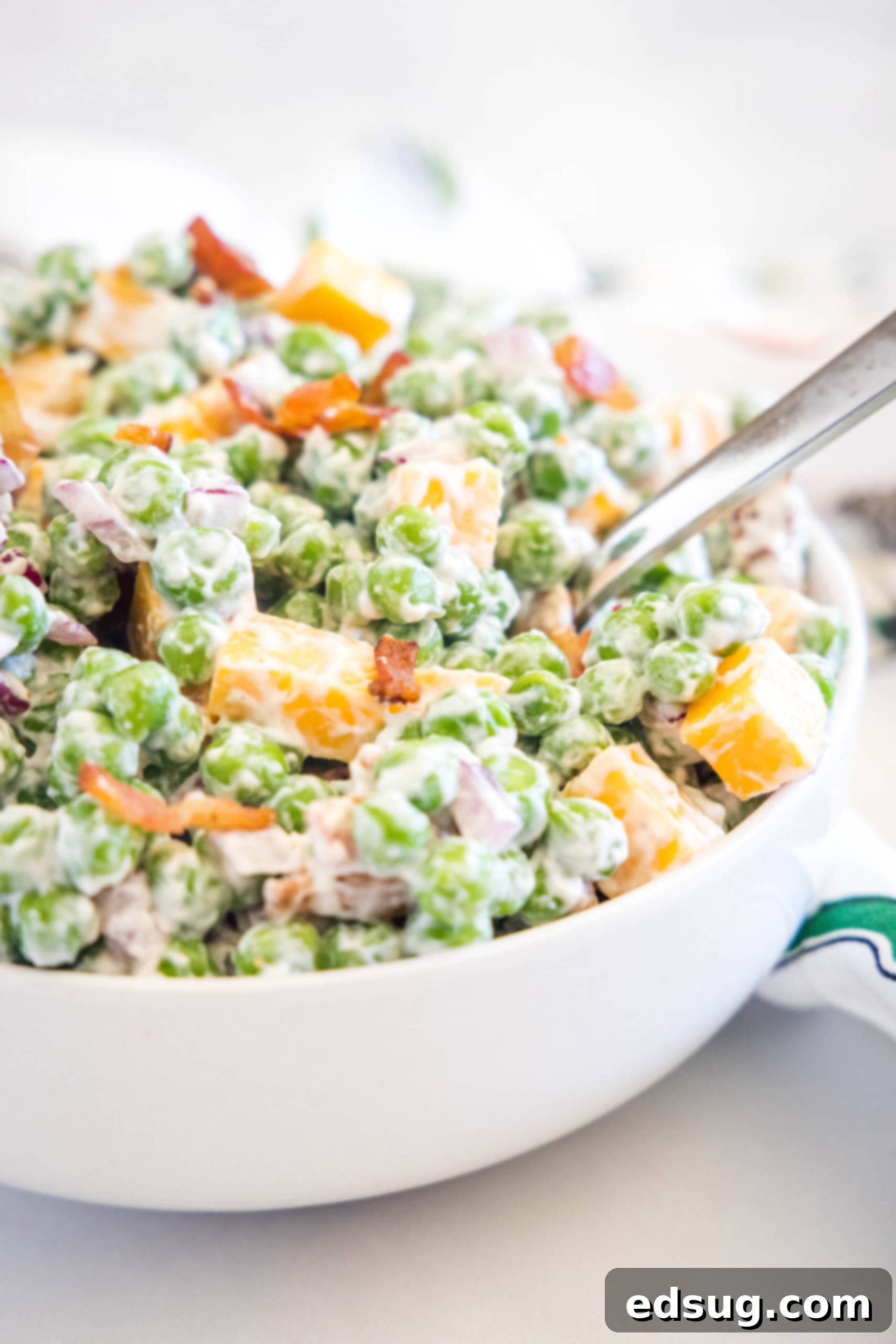 Cool and creamy pea salad is an easy side packed with crispy bacon, cheddar morsels, and red onions tossed in tangy sour cream dressing. Pea salad served in a white bowl with a spoon, ready to be enjoyed.