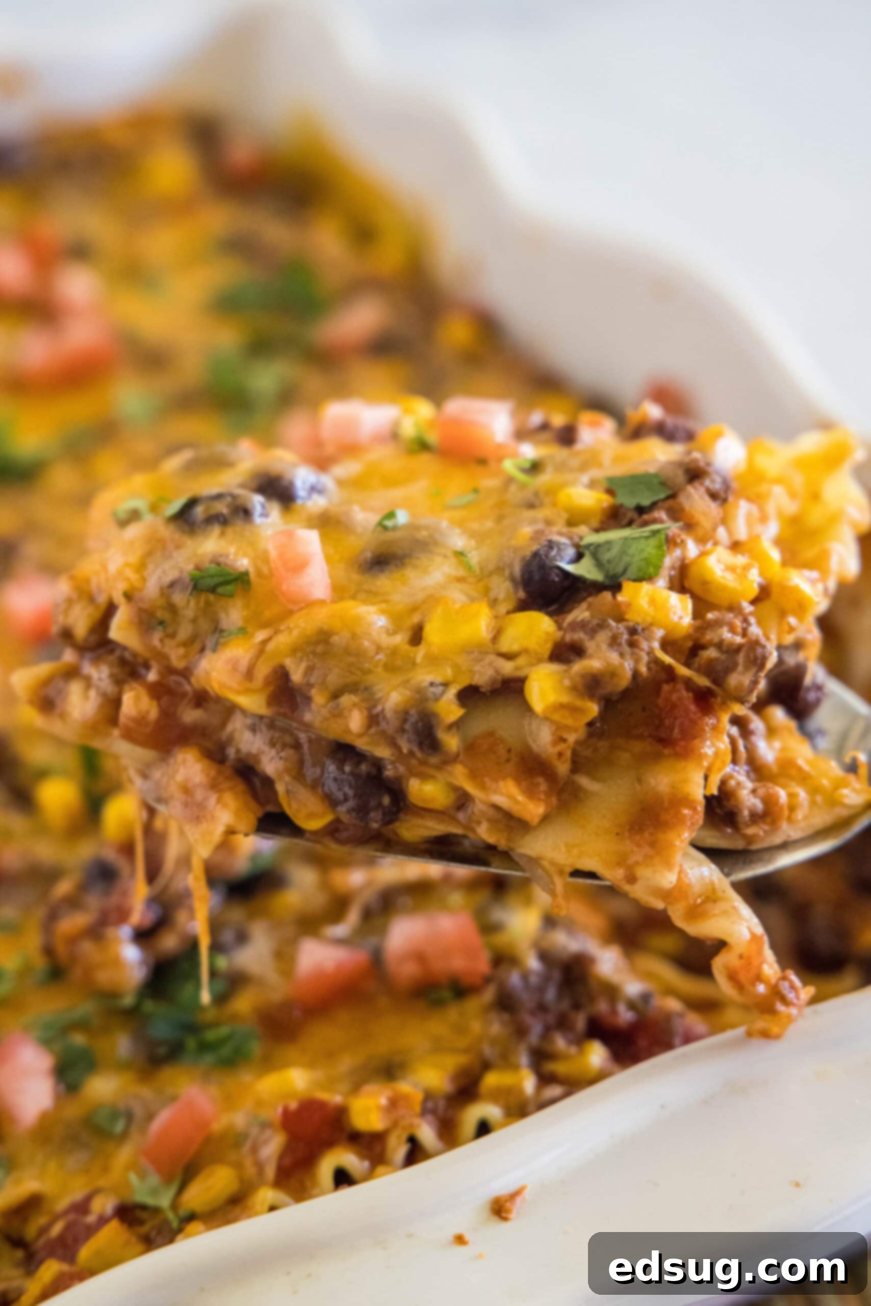 This Mexican lasagna has layers of Tex-Mex flavor, with savory beef, corn, beans, enchilada sauce, and cheese baked between lasagna noodles. A perfectly portioned slice of Mexican lasagna being lifted from a ceramic baking dish, showing off its distinct, colorful layers.