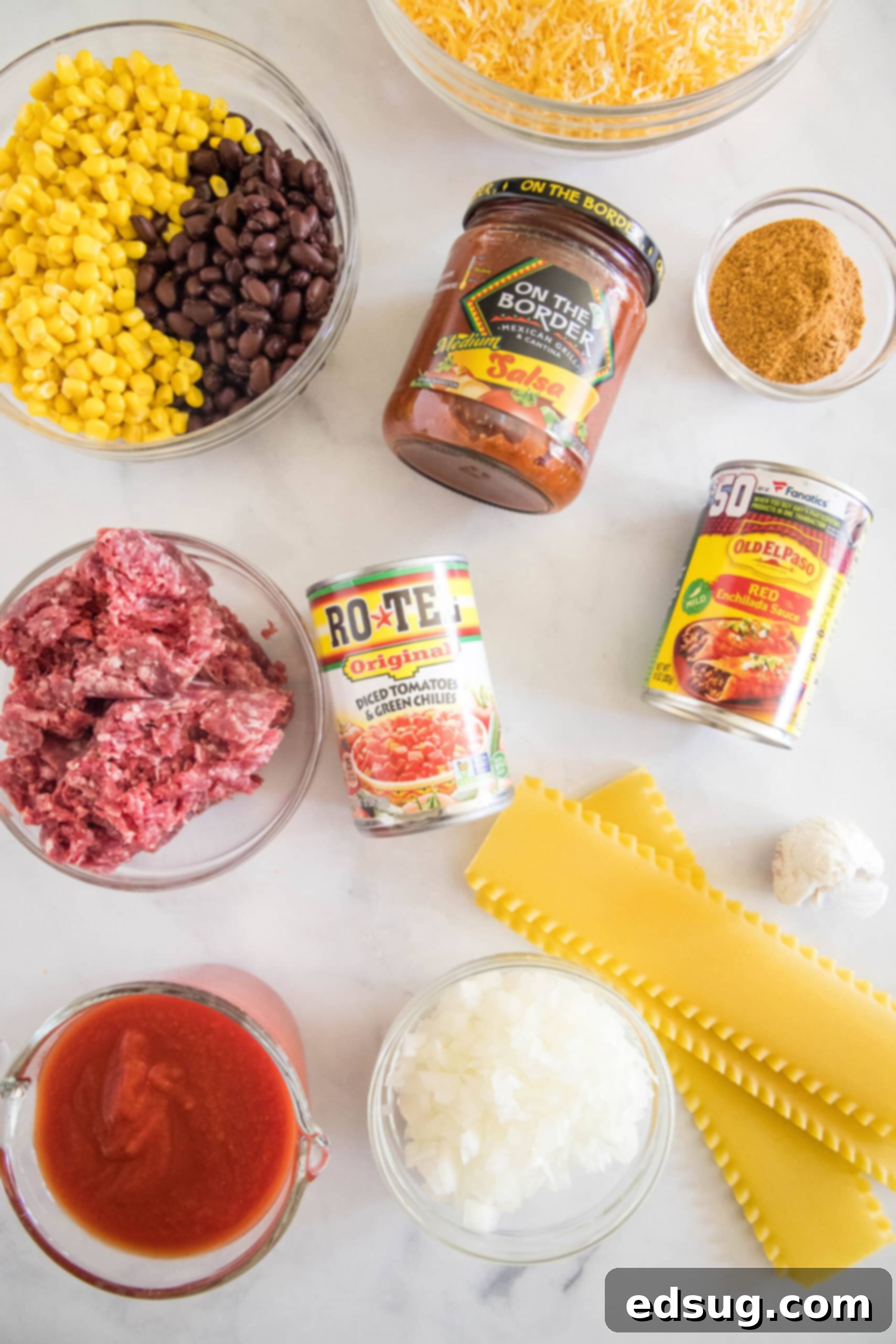 This Mexican lasagna has layers of Tex-Mex flavor, with savory beef, corn, beans, enchilada sauce, and cheese baked between lasagna noodles. Various ingredients for Mexican lasagna laid out on a white surface, including ground beef, onions, garlic, taco seasoning, canned tomatoes, black beans, corn, enchilada sauce, tomato sauce, salsa, lasagna noodles, and shredded cheese.