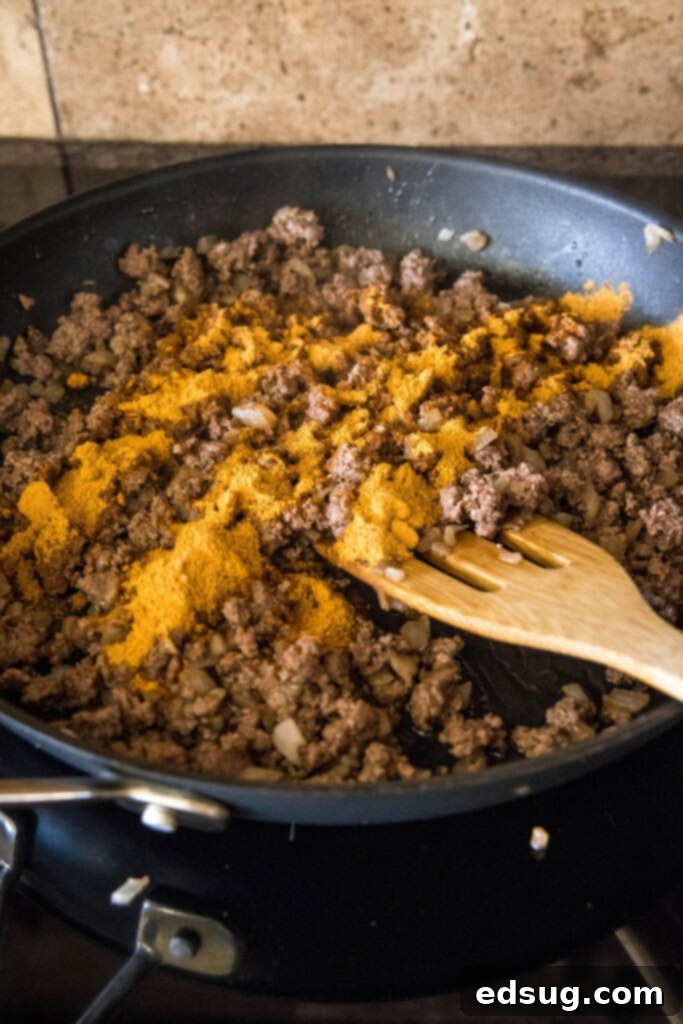This Mexican lasagna has layers of Tex-Mex flavor, with savory beef, corn, beans, enchilada sauce, and cheese baked between lasagna noodles. Browned ground beef topped with taco seasoning in a skillet with a wooden spatula, ready for further ingredients.