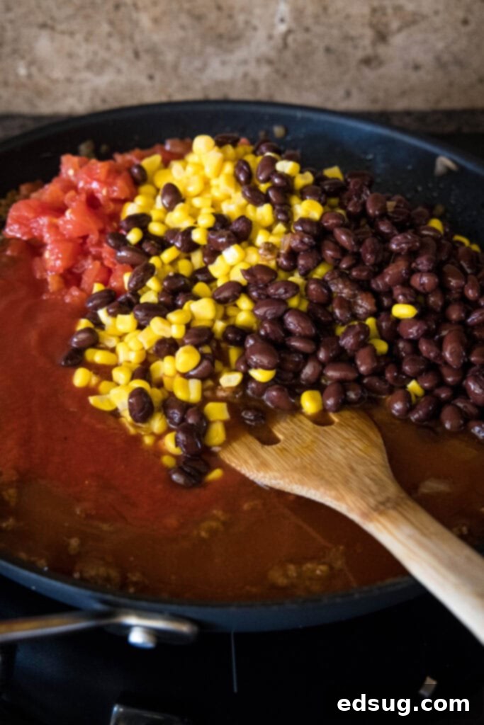 This Mexican lasagna has layers of Tex-Mex flavor, with savory beef, corn, beans, enchilada sauce, and cheese baked between lasagna noodles. A wooden spatula stirs black beans, corn, and diced tomatoes into a skillet with the ground beef lasagna filling, showcasing the vibrant colors of the ingredients.