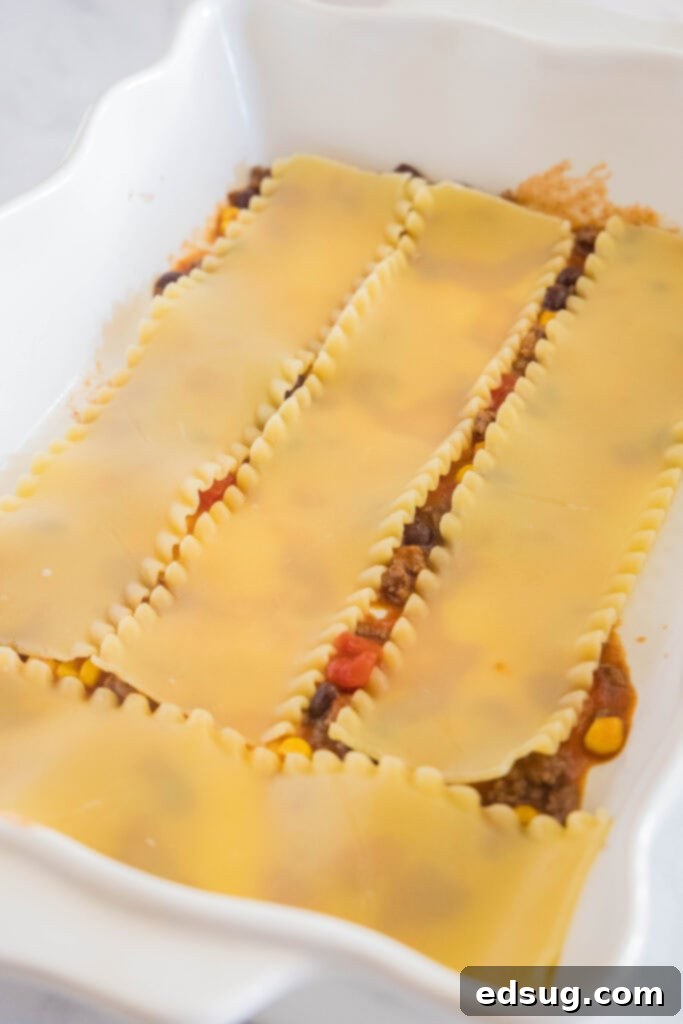 This Mexican lasagna has layers of Tex-Mex flavor, with savory beef, corn, beans, enchilada sauce, and cheese baked between lasagna noodles. Lasagna noodles layered over a rich Mexican ground beef filling in a white ceramic baking dish, showing the initial assembly.