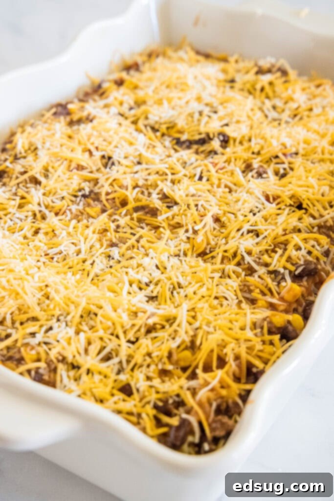 This Mexican lasagna has layers of Tex-Mex flavor, with savory beef, corn, beans, enchilada sauce, and cheese baked between lasagna noodles. Assembled Mexican lasagna, abundantly topped with shredded cheese, waiting to be baked in a white ceramic baking dish.