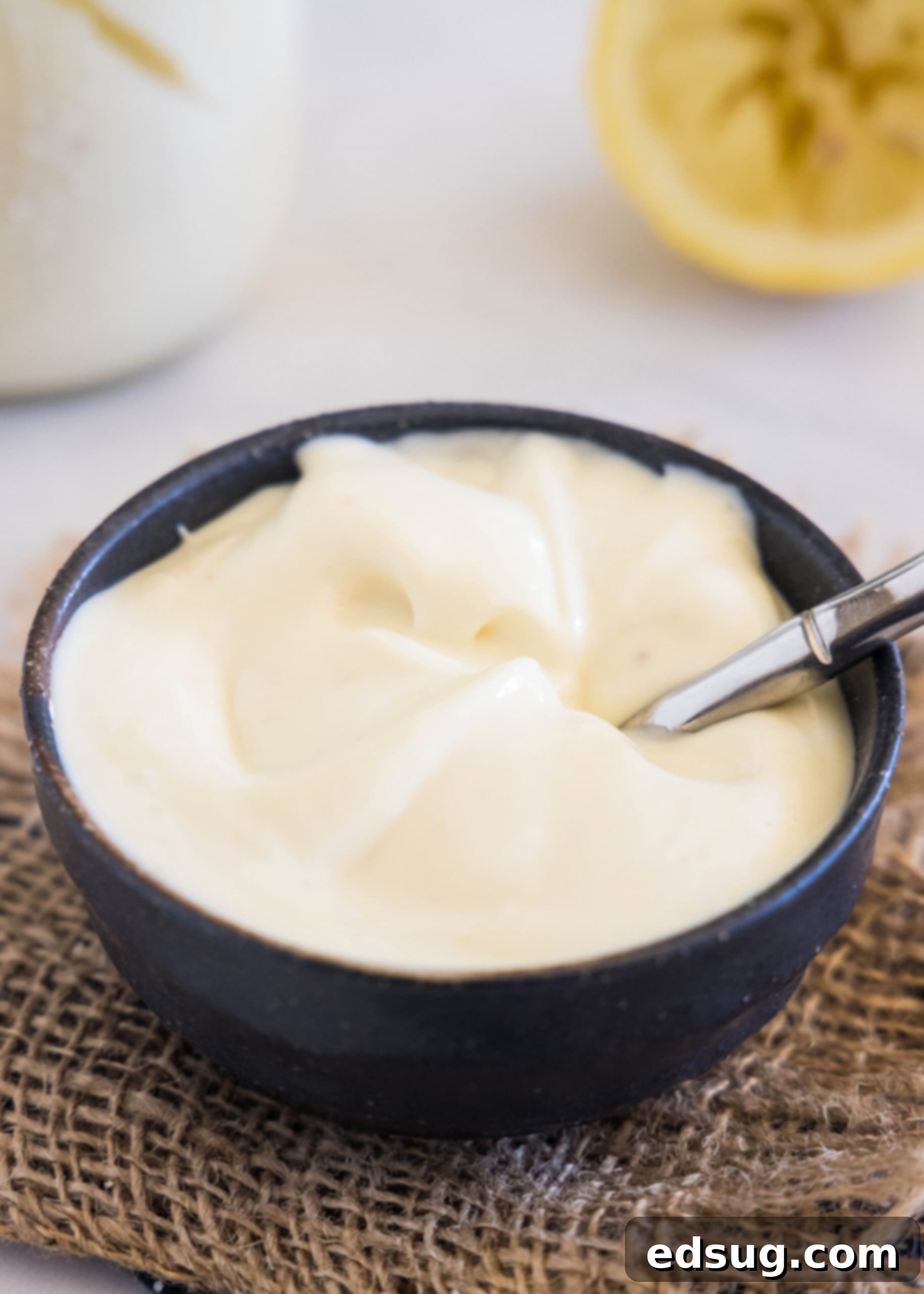 Rich, creamy homemade mayonnaise in a black bowl with a spoon, accompanied by a freshly juiced lemon and a jar of homemade mayonnaise in the background, highlighting its fresh appeal.