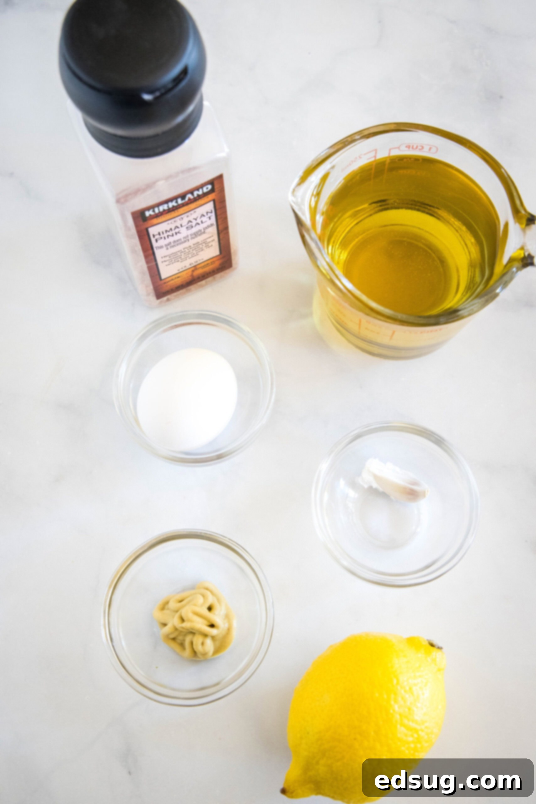 A collection of fresh ingredients laid out on a countertop, including a whole egg, a jar of Dijon mustard, a lemon cut in half, garlic cloves, and a bottle of avocado oil, ready for making homemade mayo.