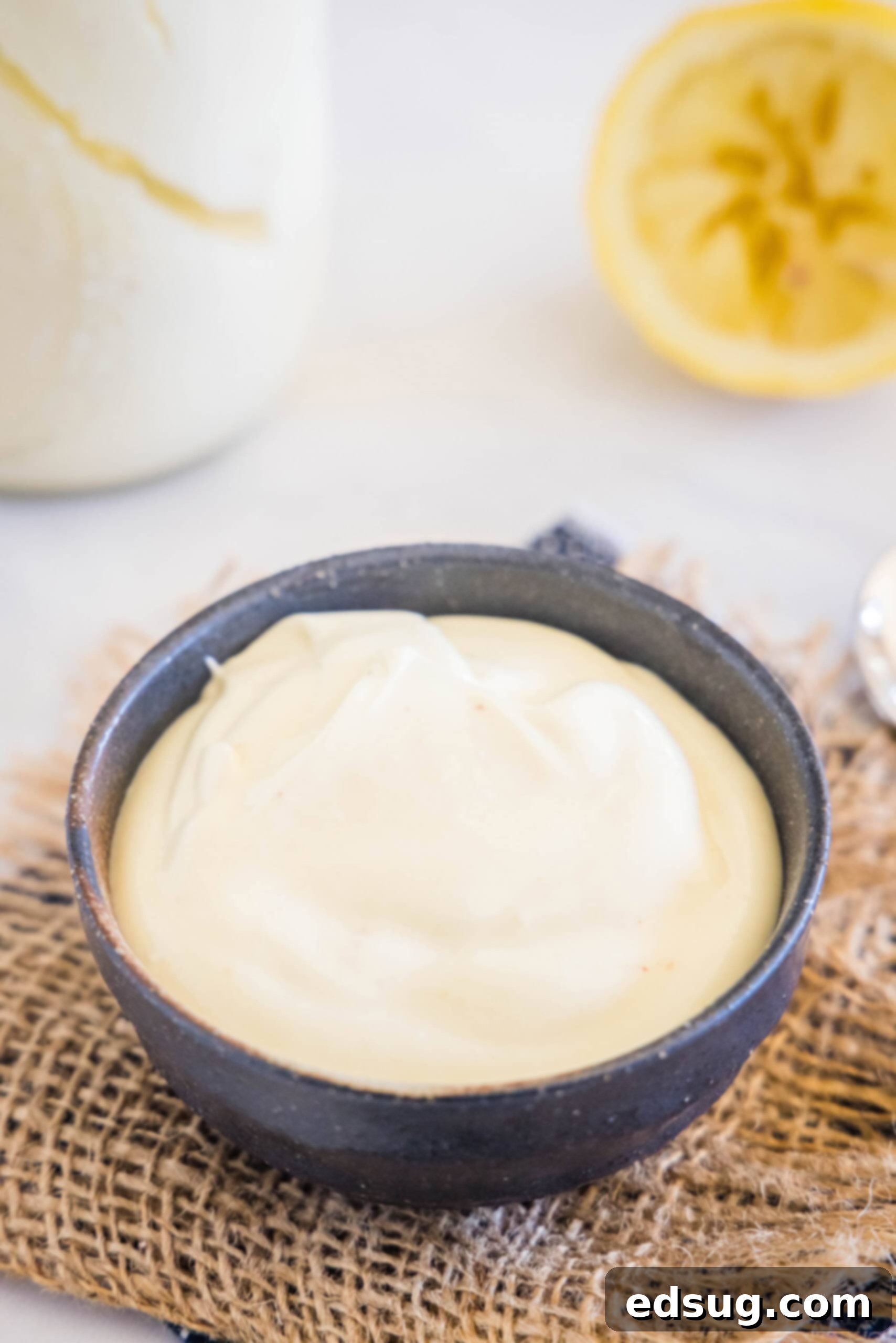 Creamy homemade mayonnaise presented in a sleek black bowl, with a juiced lemon half and another jar of mayo subtly blurred in the background, emphasizing its freshness.