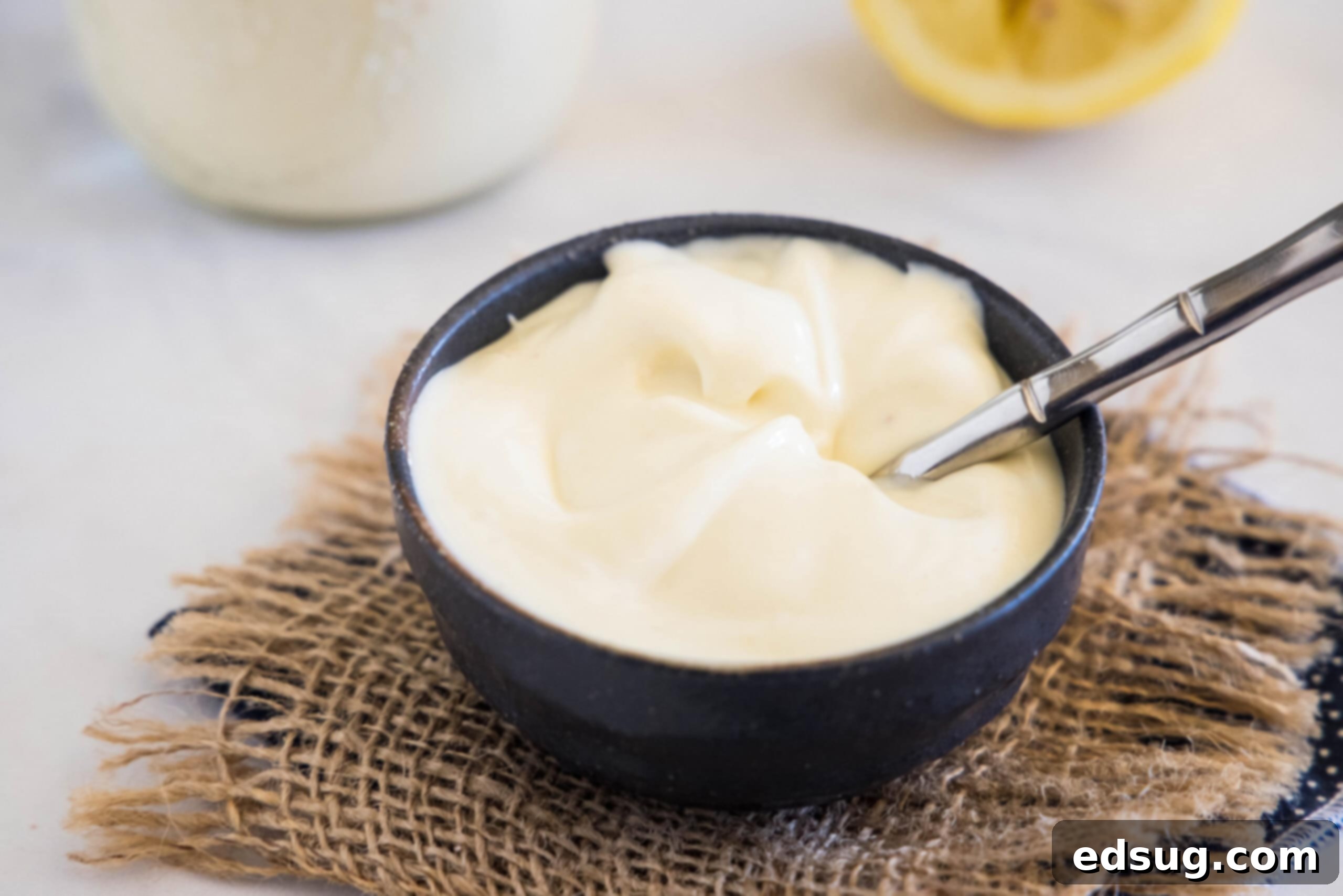 A black bowl filled with creamy homemade mayonnaise, with a spoon resting in it, alongside a freshly squeezed lemon and a jar of mayo, creating an appealing kitchen scene.