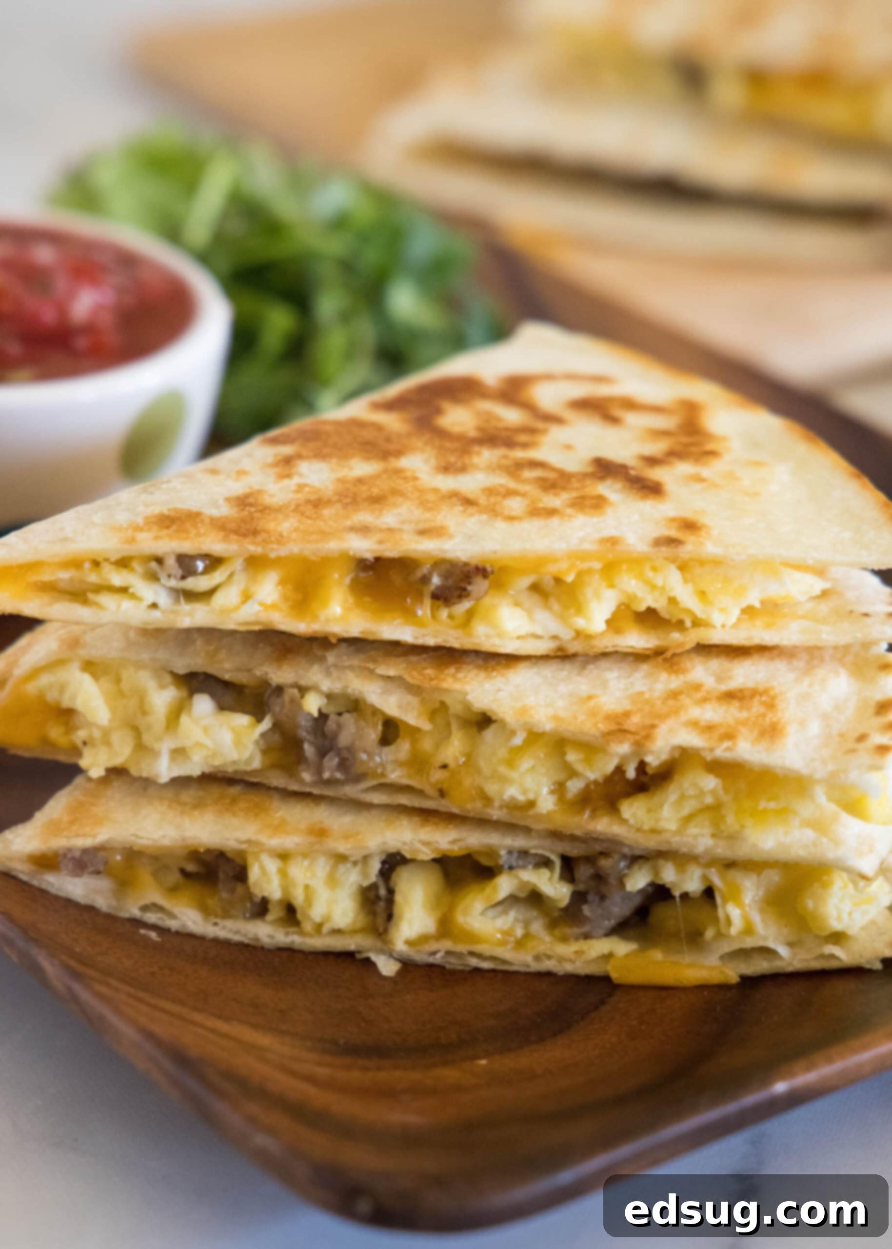 Rise and shine with this easy breakfast quesadilla recipe! Made with eggs, savory sausage, and melty cheese folded between warm tortillas. Three golden-brown breakfast quesadillas stacked on a rustic wooden platter, with a bowl of fresh salsa in the background.