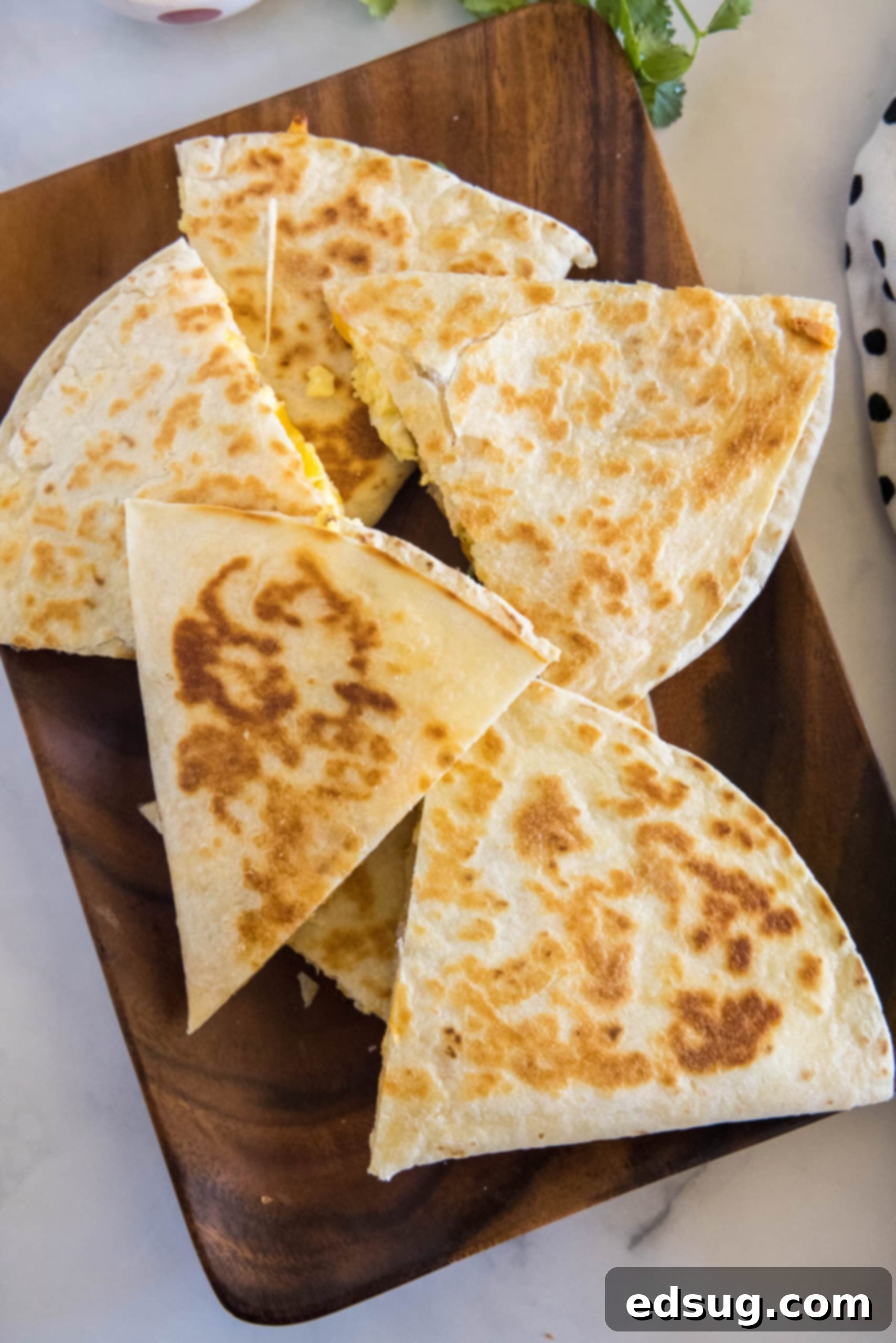 Rise and shine with this easy breakfast quesadilla recipe! Made with eggs, savory sausage, and melty cheese folded between warm tortillas. Overhead view of perfectly cooked breakfast quesadillas, stacked on a wooden serving platter, garnished and ready.