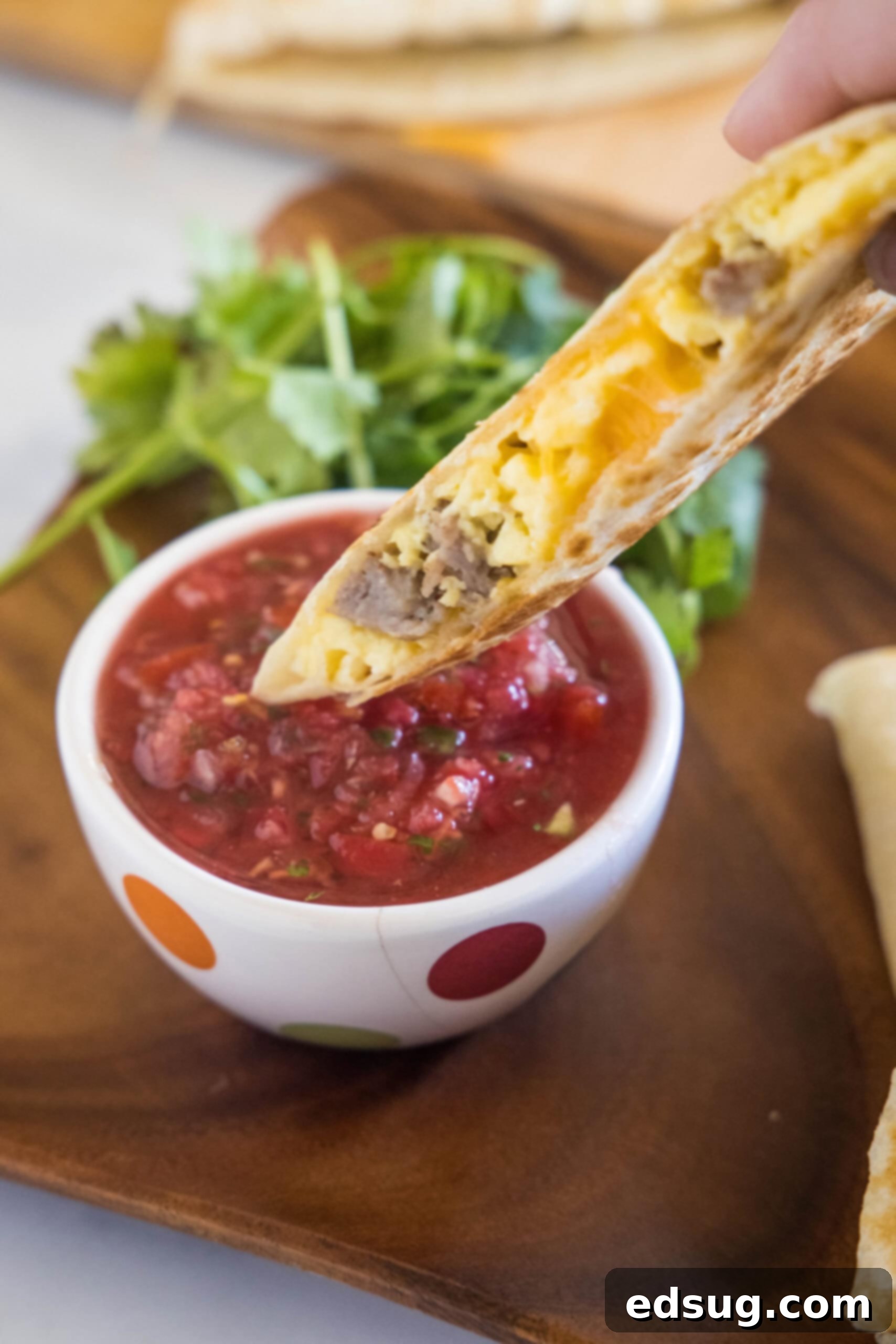 Rise and shine with this easy breakfast quesadilla recipe! Made with eggs, savory sausage, and melty cheese folded between warm tortillas. A breakfast quesadilla wedge being dipped into a bowl of vibrant red salsa.
