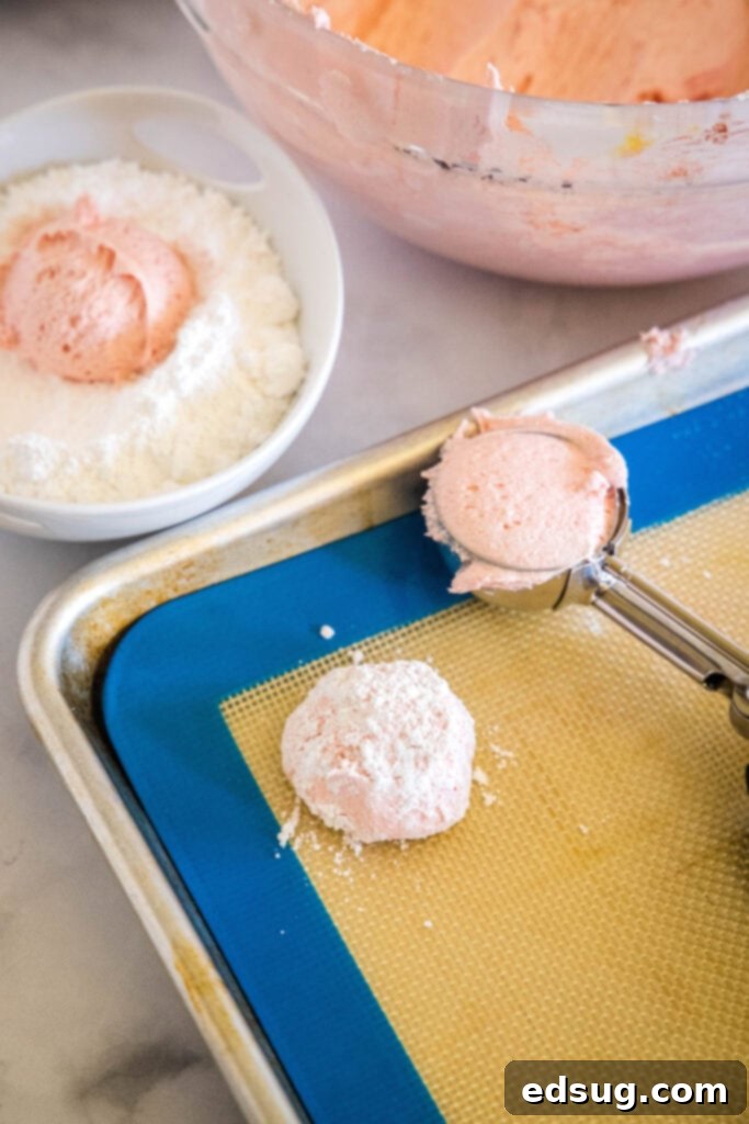 A cookie scoop resting on a lined baking sheet next to a portioned Cool Whip cookie dough ball, perfectly rolled in powdered sugar and ready for baking. A bowl of dough is in the background.