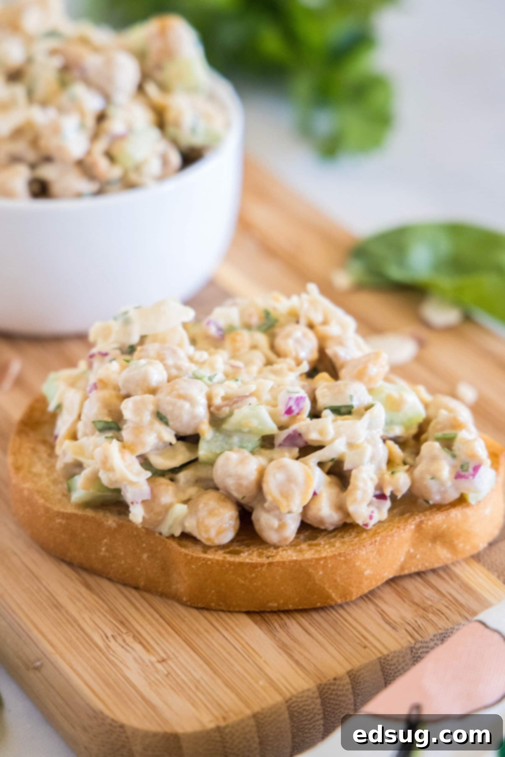 Creamy chickpea chicken salad served on a slice of rustic bread on a wooden cutting board, with a bowl of the salad in the background, garnished with fresh parsley.
