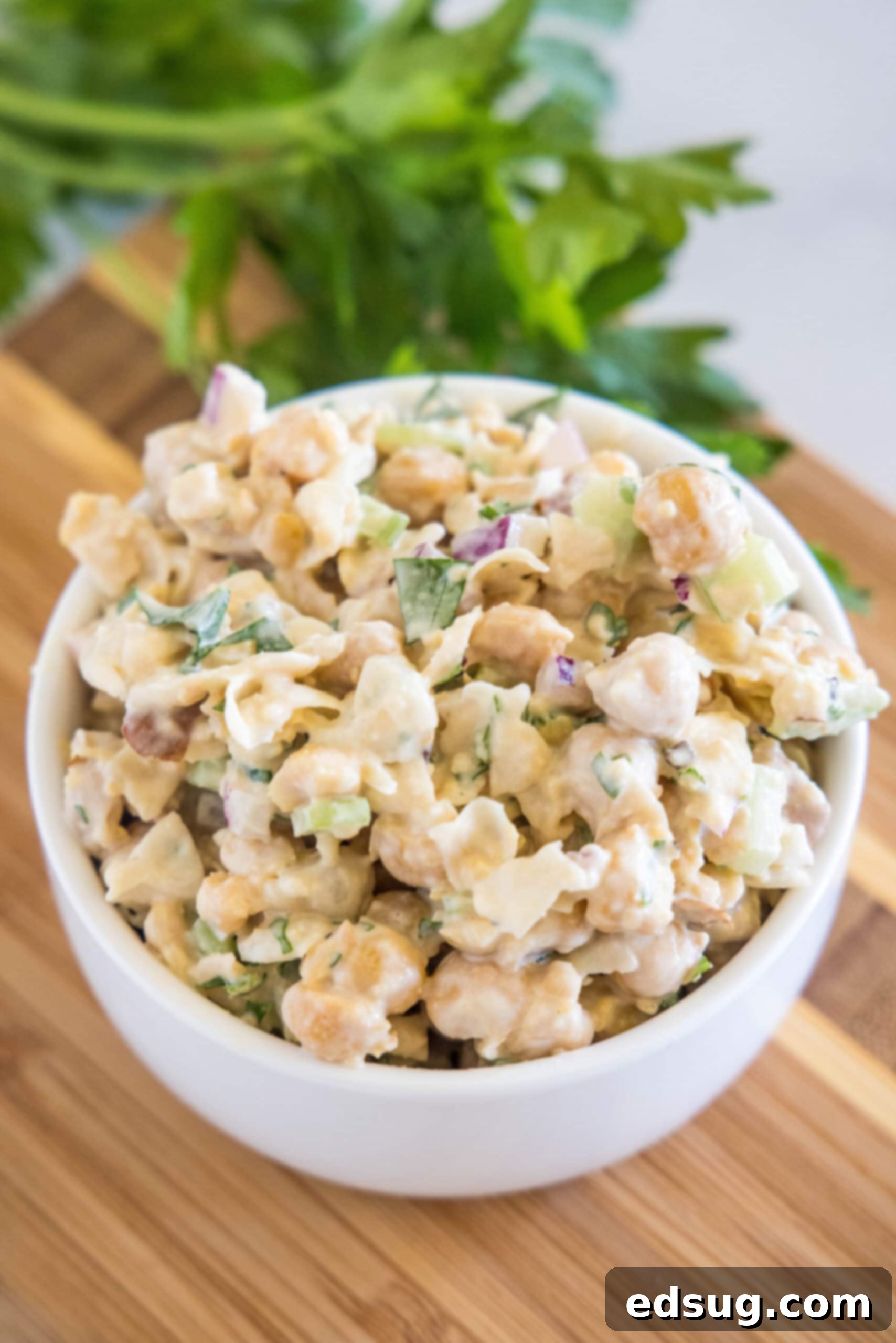 A large glass bowl filled with freshly made chickpea chicken salad, garnished with fresh herbs, resting on a rustic wooden cutting board.