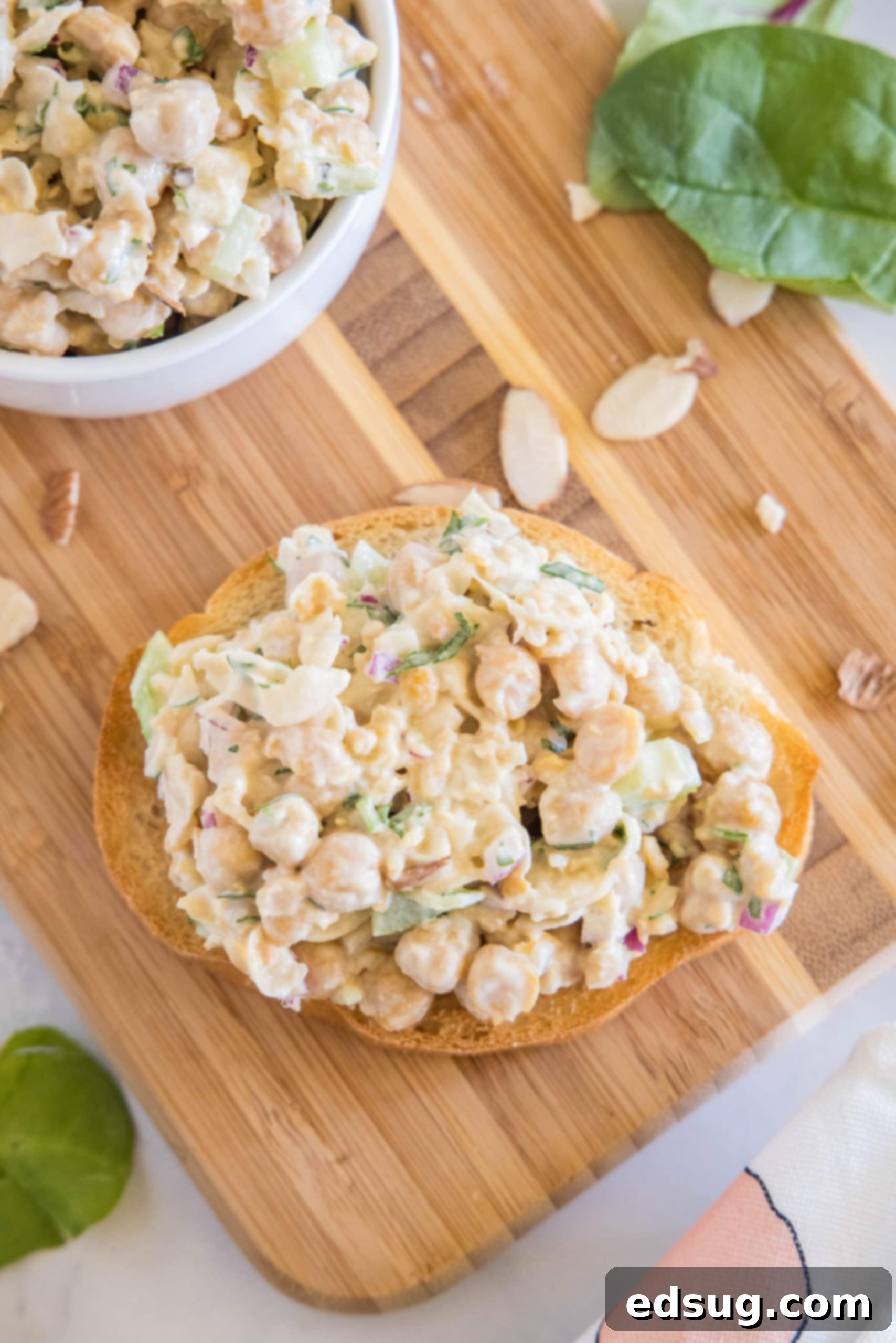 Overhead shot of chickpea chicken salad generously spread on a slice of toasted bread, with a full bowl of salad beside it, all on a wooden cutting board.