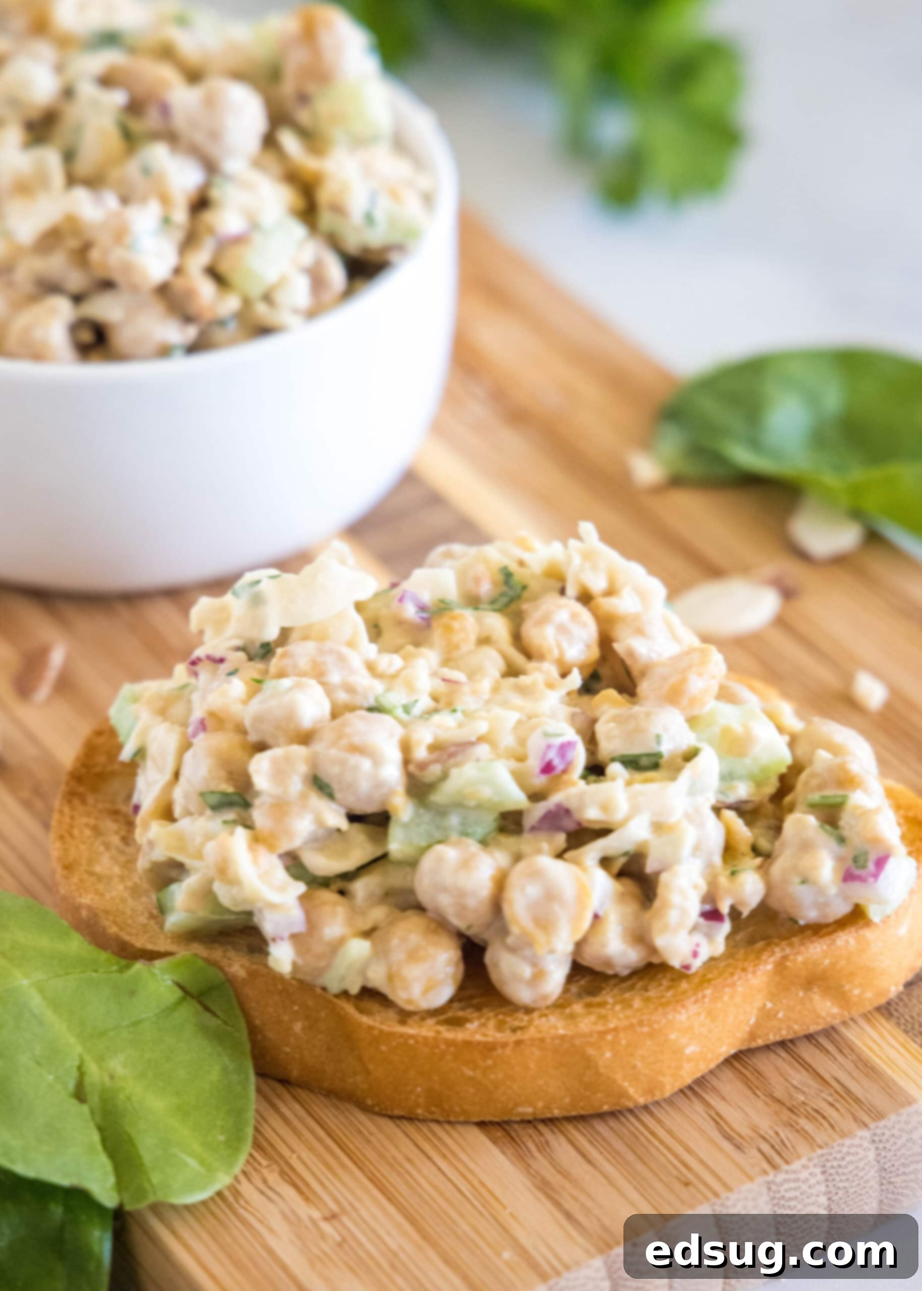 Chickpea chicken salad elegantly served on a thick slice of artisanal bread on a wooden cutting board, with a large bowl of the salad subtly blurred in the background.