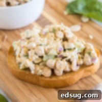 Chickpea chicken salad served on a slice of bread on a wooden cutting board, with a bowl of chickpea salad in the background.